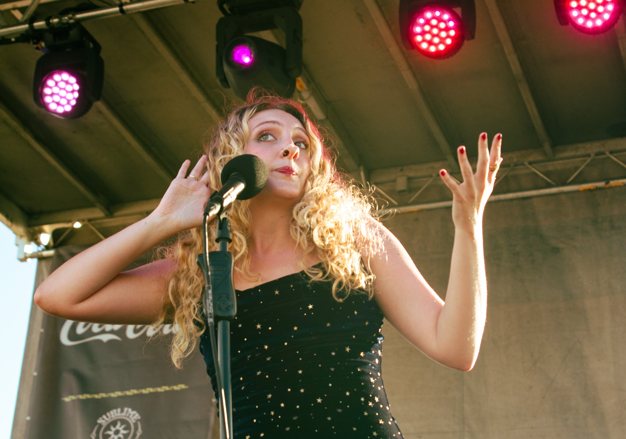 A young woman with curly blonde hair performs on a stage with purple and red stage lights.