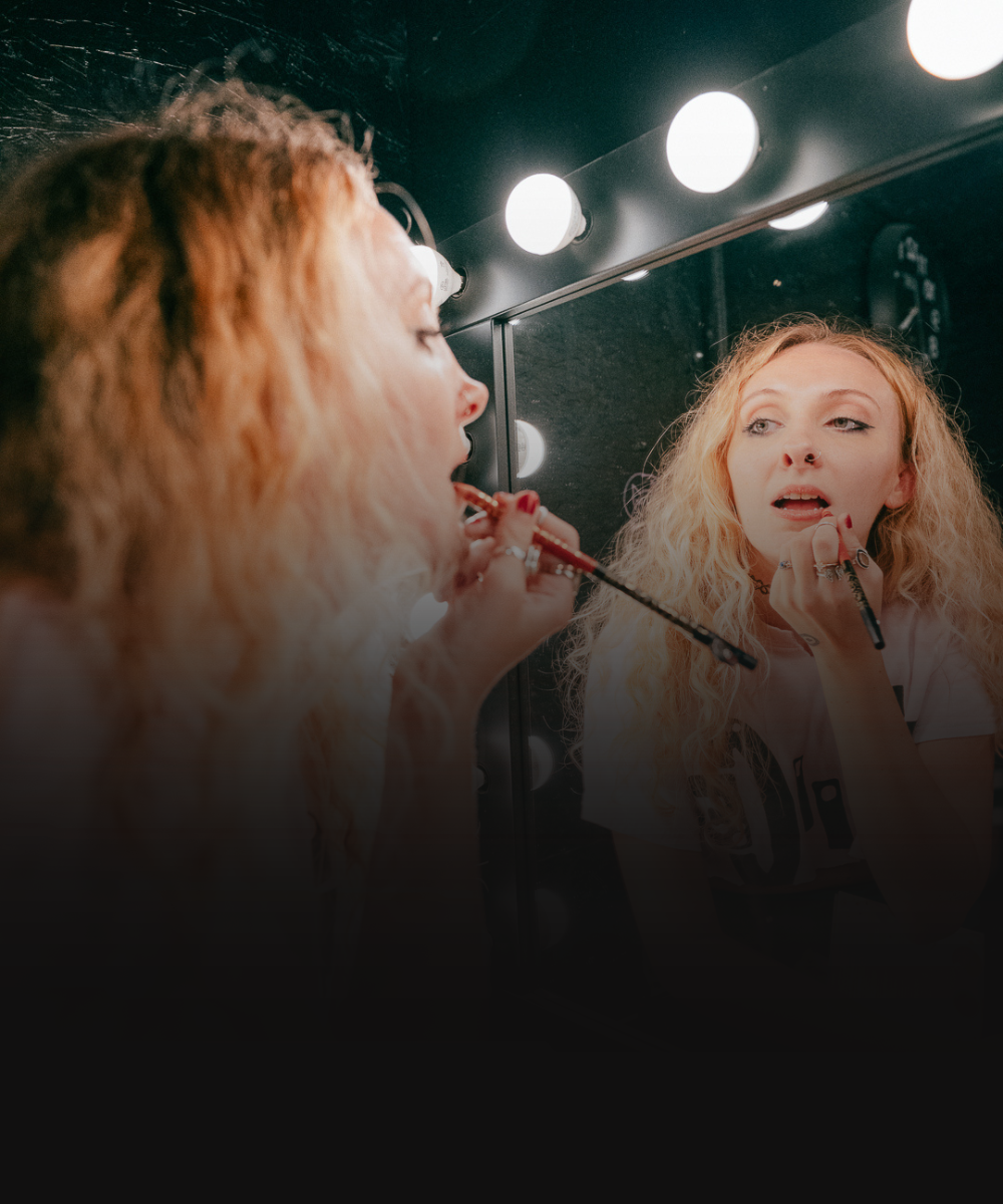 A young woman with curly blonde hair applying red lipliner in front of a mirror illuminated by round vanity lights.
