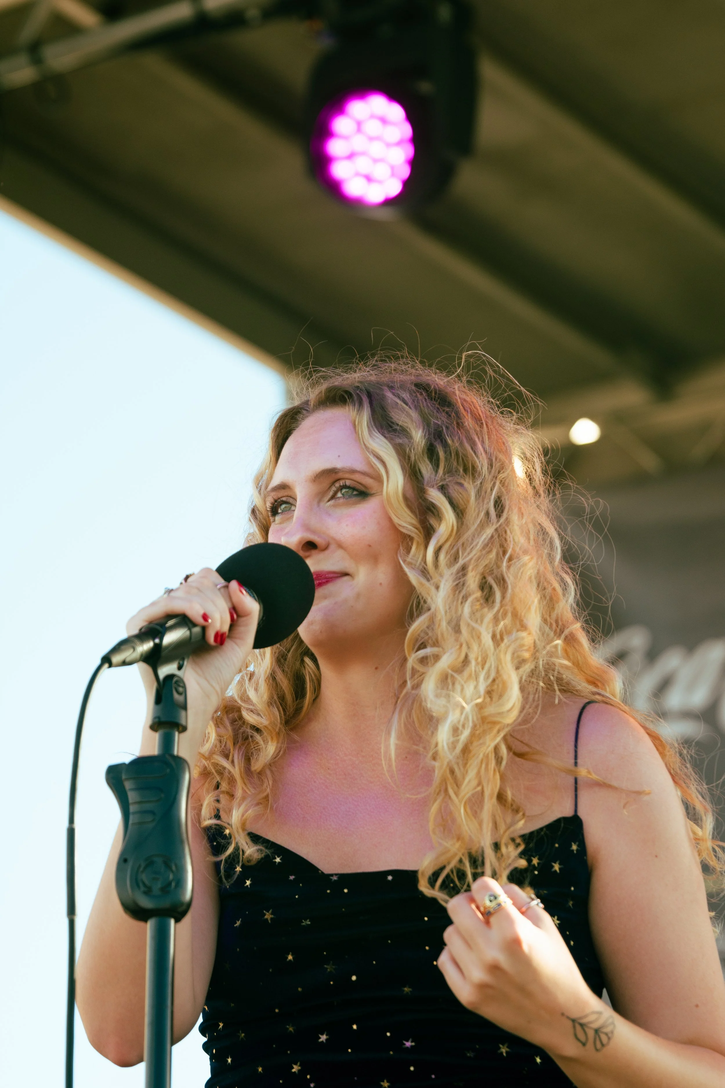 A woman with long curly blonde hair holding a microphone and singing on stage, under a purple stage light.