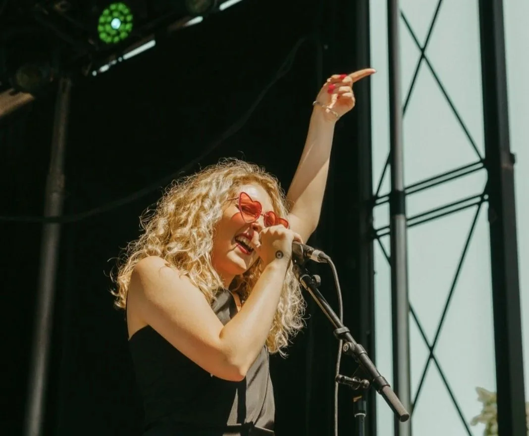 A woman performing on stage, wearing red heart-shaped sunglasses, singing into a microphone, with one arm raised.