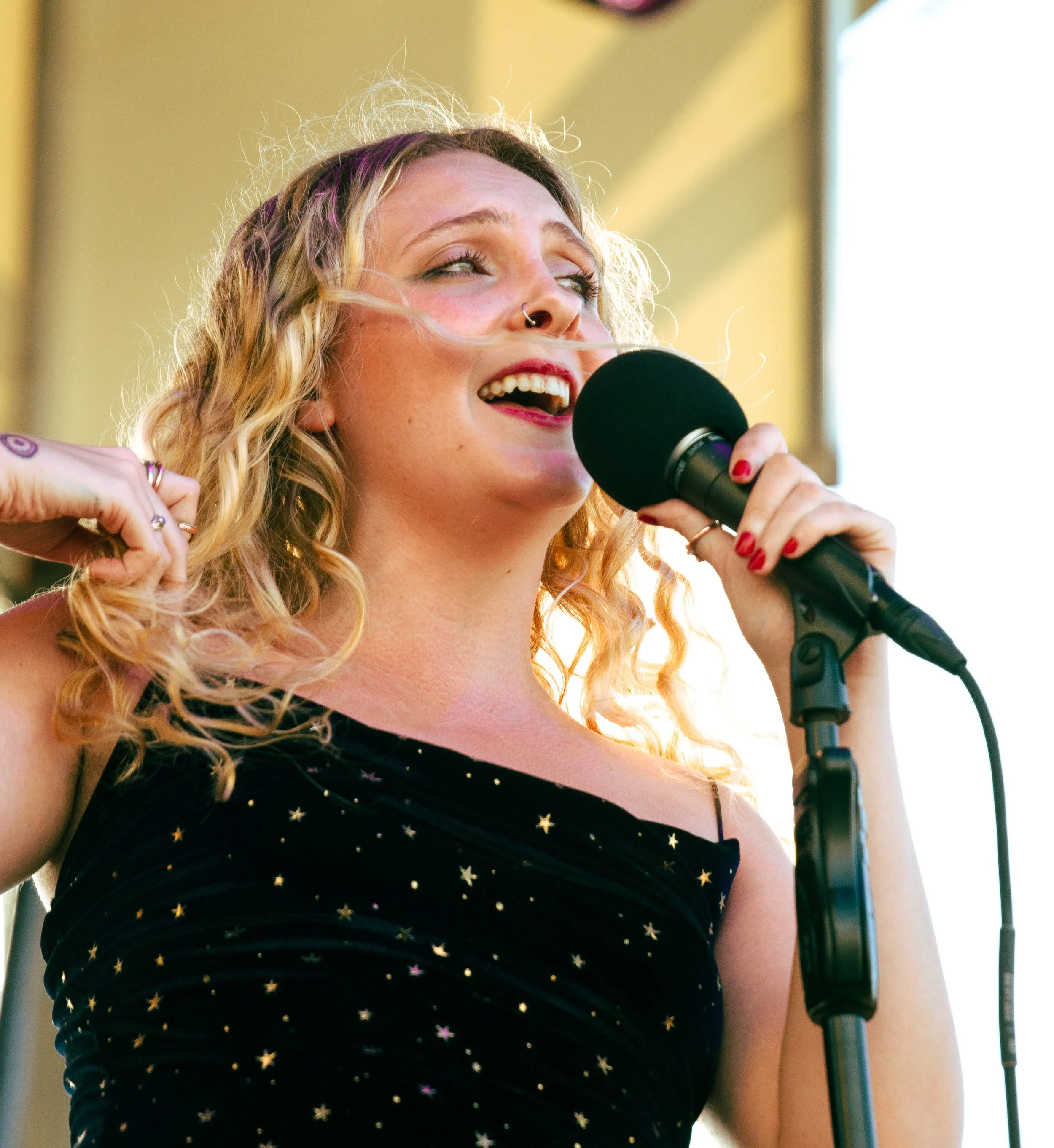 Close-up of a young woman with curly blonde hair, holding a microphone, singing passionately, wearing a black dress with gold star patterns.