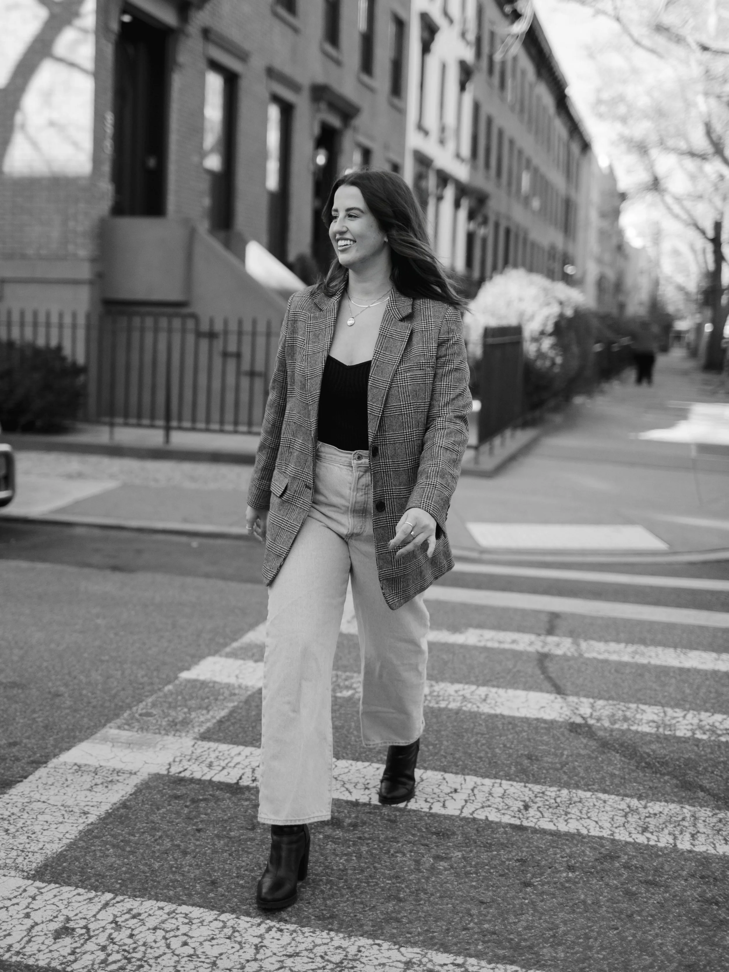 A woman with dark hair smiling, walking across a crosswalk in an urban neighborhood. She is wearing a plaid blazer, a black top, high-waisted jeans, and black boots. The background shows residential buildings, trees, and other pedestrians.