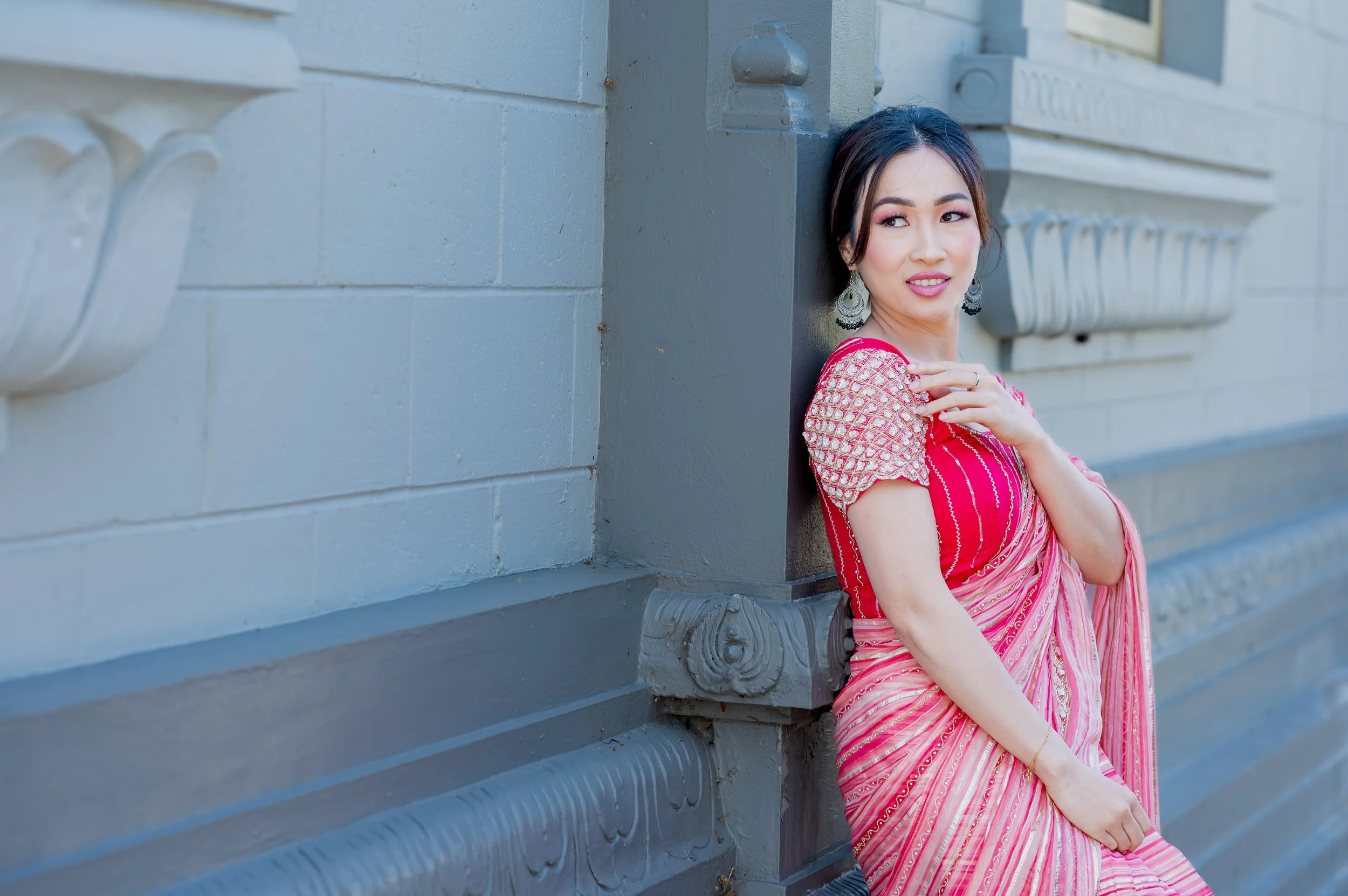 A woman wearing a red and pink traditional Indian saree with earrings is leaning against a gray building wall, looking to her right with a slight smile, outdoors during daytime.