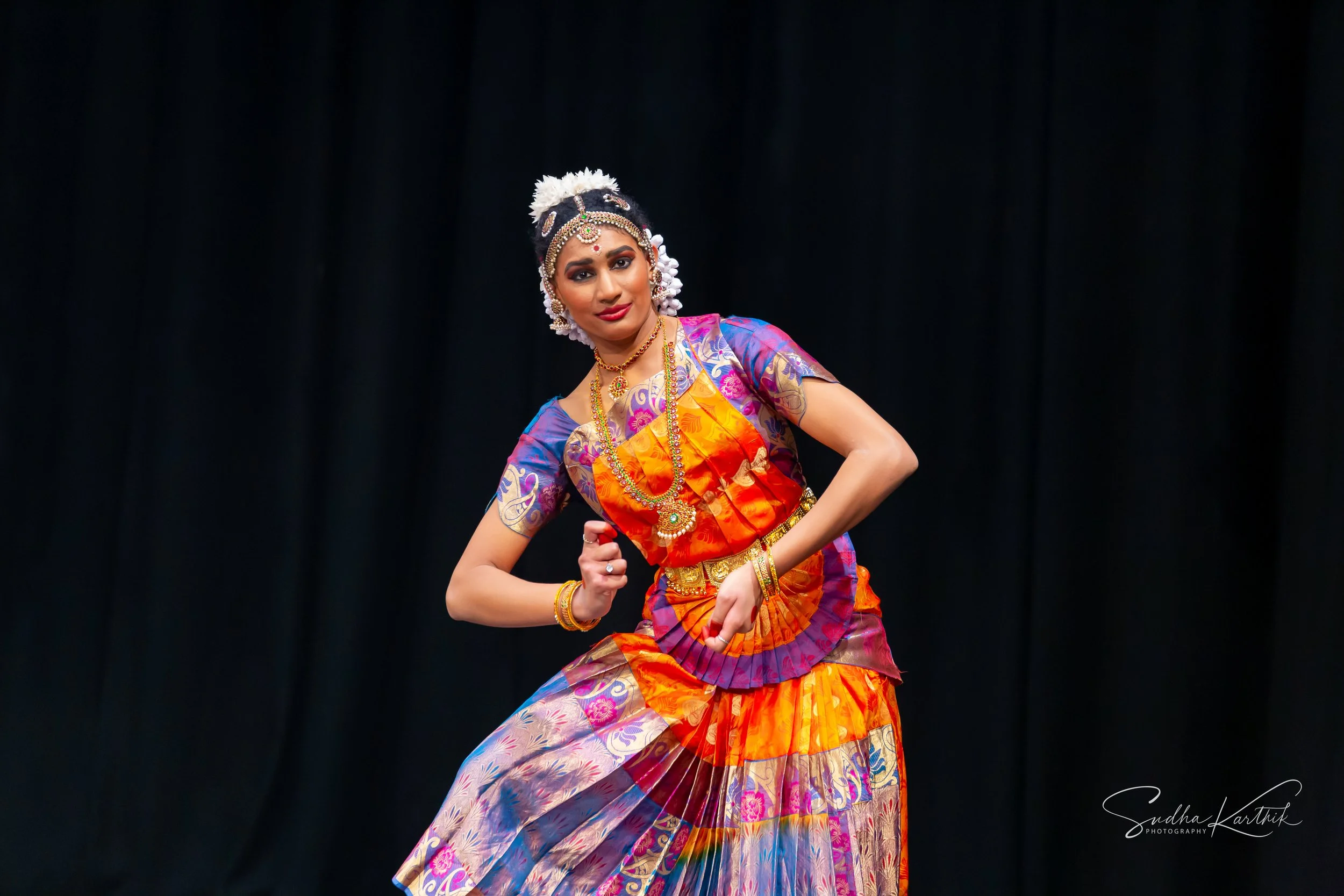 A woman dressed in traditional Indian dance attire, wearing colorful silk garments and ornate jewelry, performing a classical dance pose against a black backdrop.