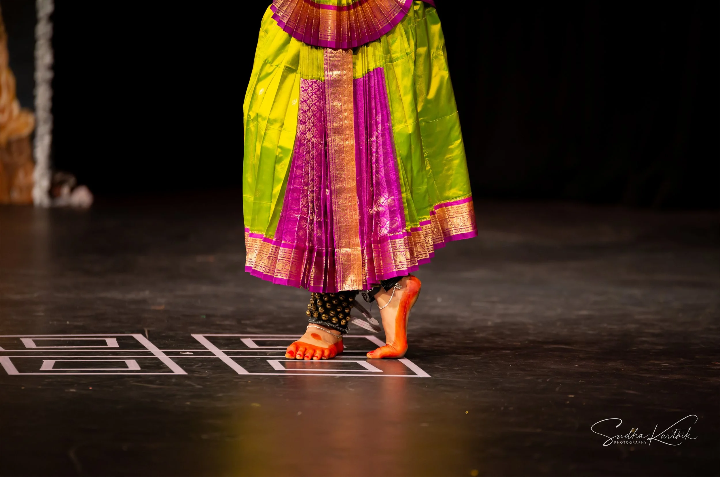 A dancer on stage wearing vibrant, traditional Indian clothing including a bright yellow skirt with pink and gold accents and ornate jewelry, with only their legs and feet visible.