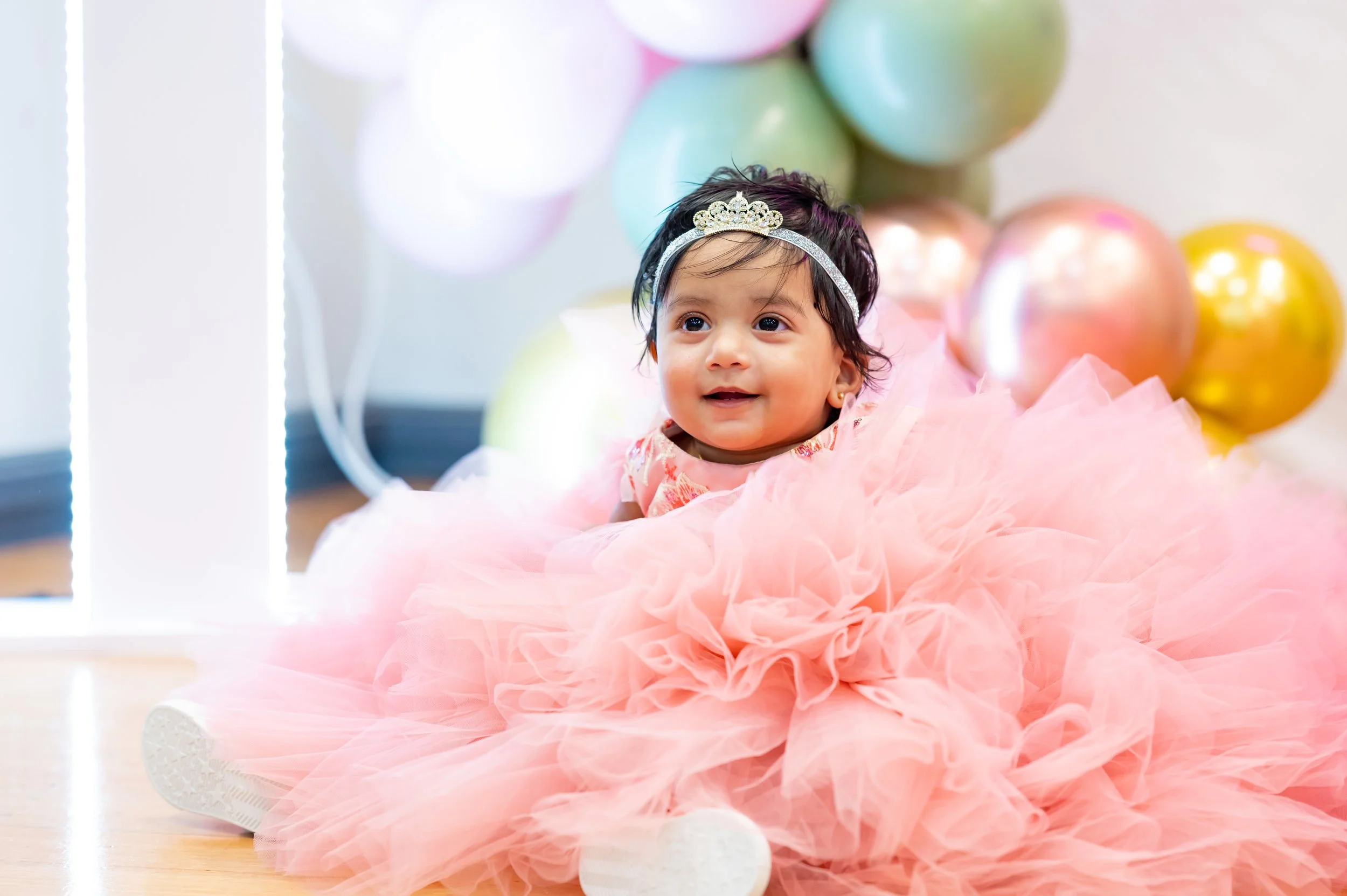 A young girl wearing a tiara and a pink tutu skirt, sitting on the floor surrounded by pastel-colored balloons.