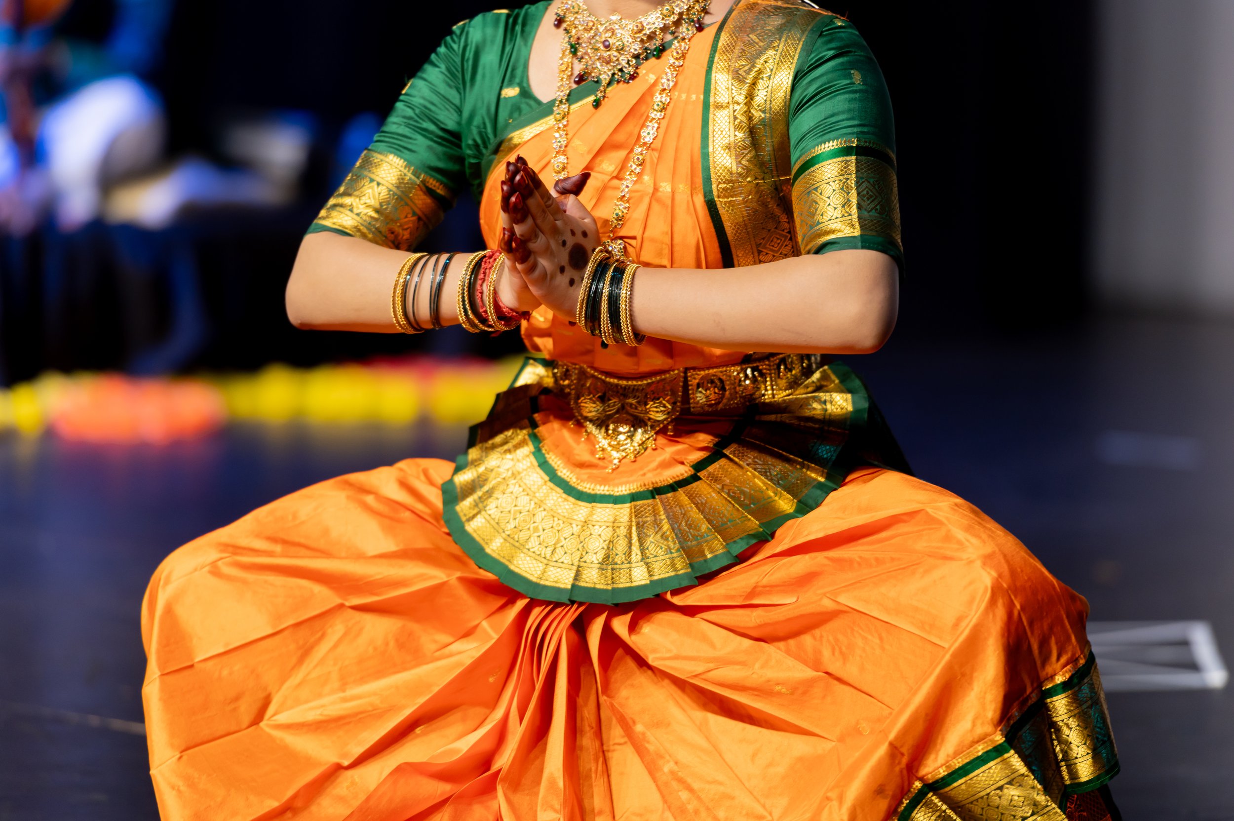 A woman dressed in traditional Indian attire, wearing an orange and green sari with gold accents, sitting in a cross-legged position with hands in a prayer gesture, adorned with jewelry and bangles.