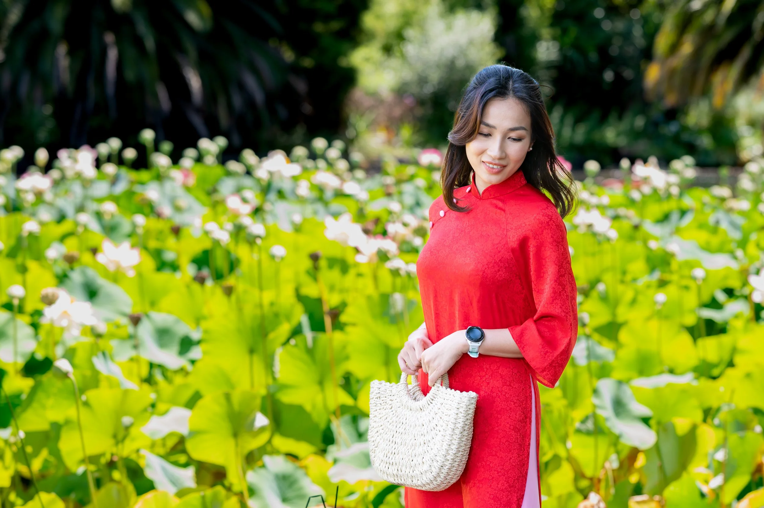A young woman wearing a red traditional dress stands outdoors in a lush green garden with blooming white flowers. She is smiling gently and holding a white woven bag, with a smartwatch on her wrist.