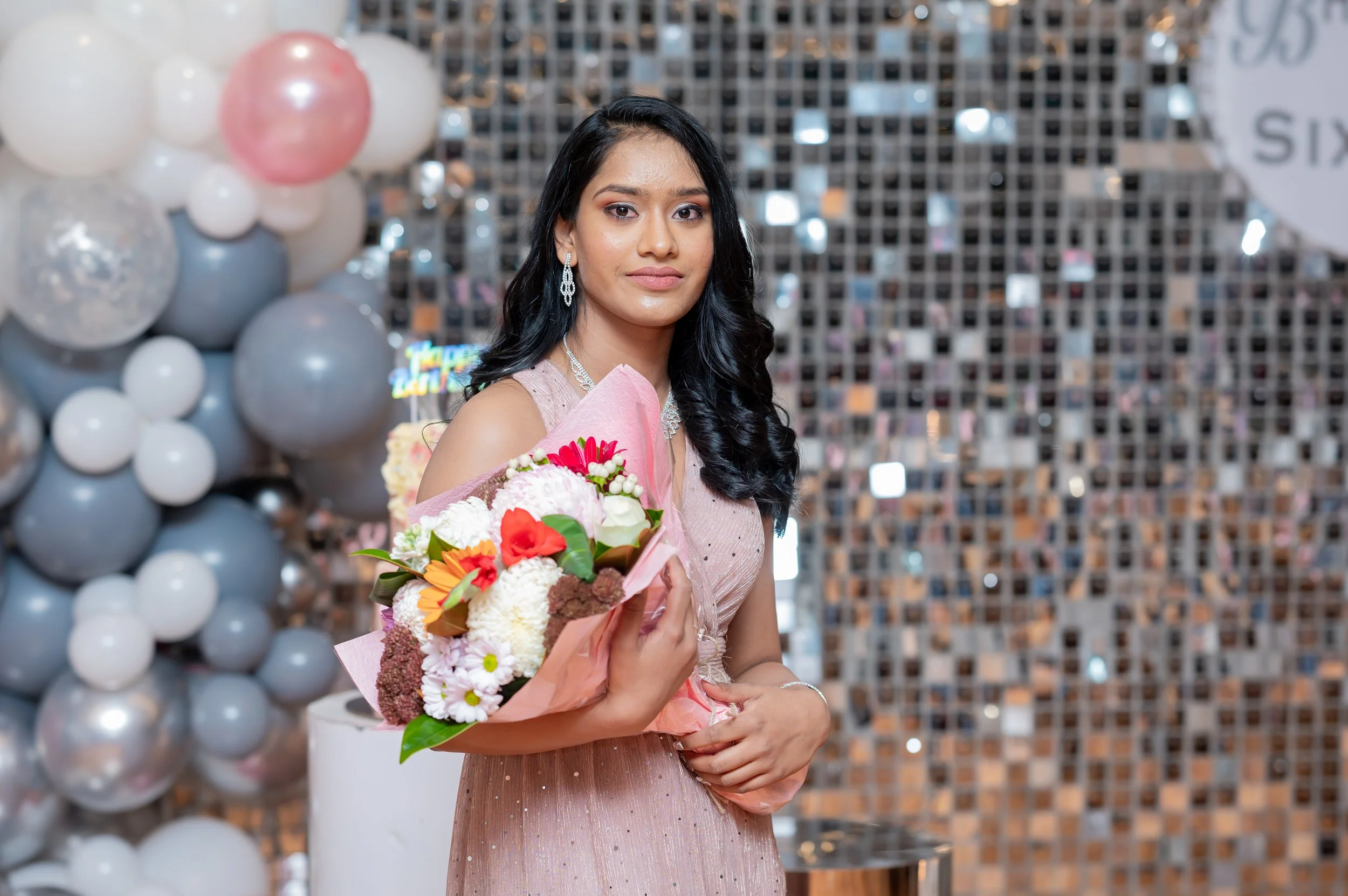 A young woman with long black wavy hair in a sleeveless pink dress holds a bouquet of colorful flowers at a celebration or event, standing in front of a backdrop with balloons and decorative tiles.