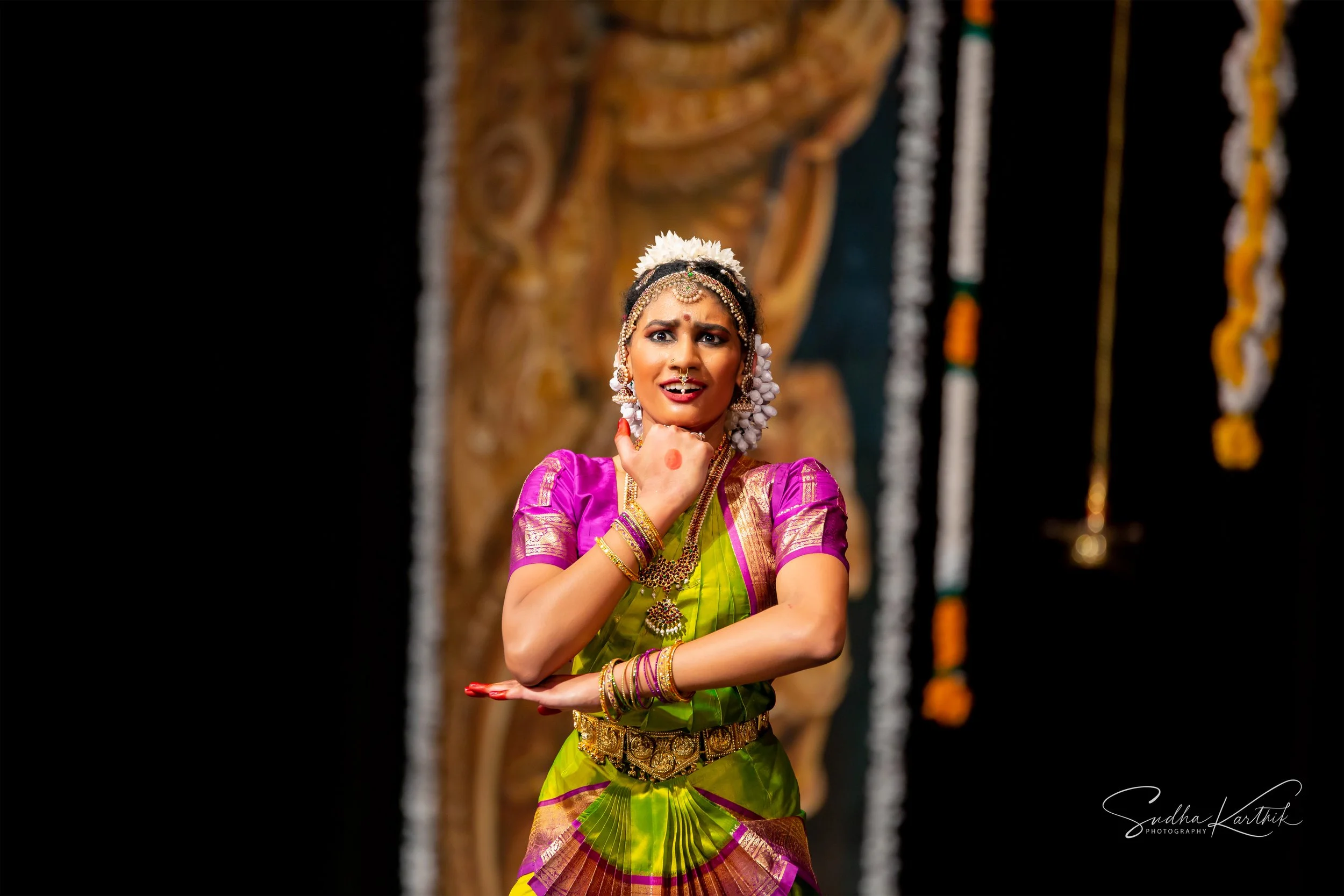 A woman dressed in traditional Indian attire performing a classical dance on stage, with ornate jewelry and a decorated background.