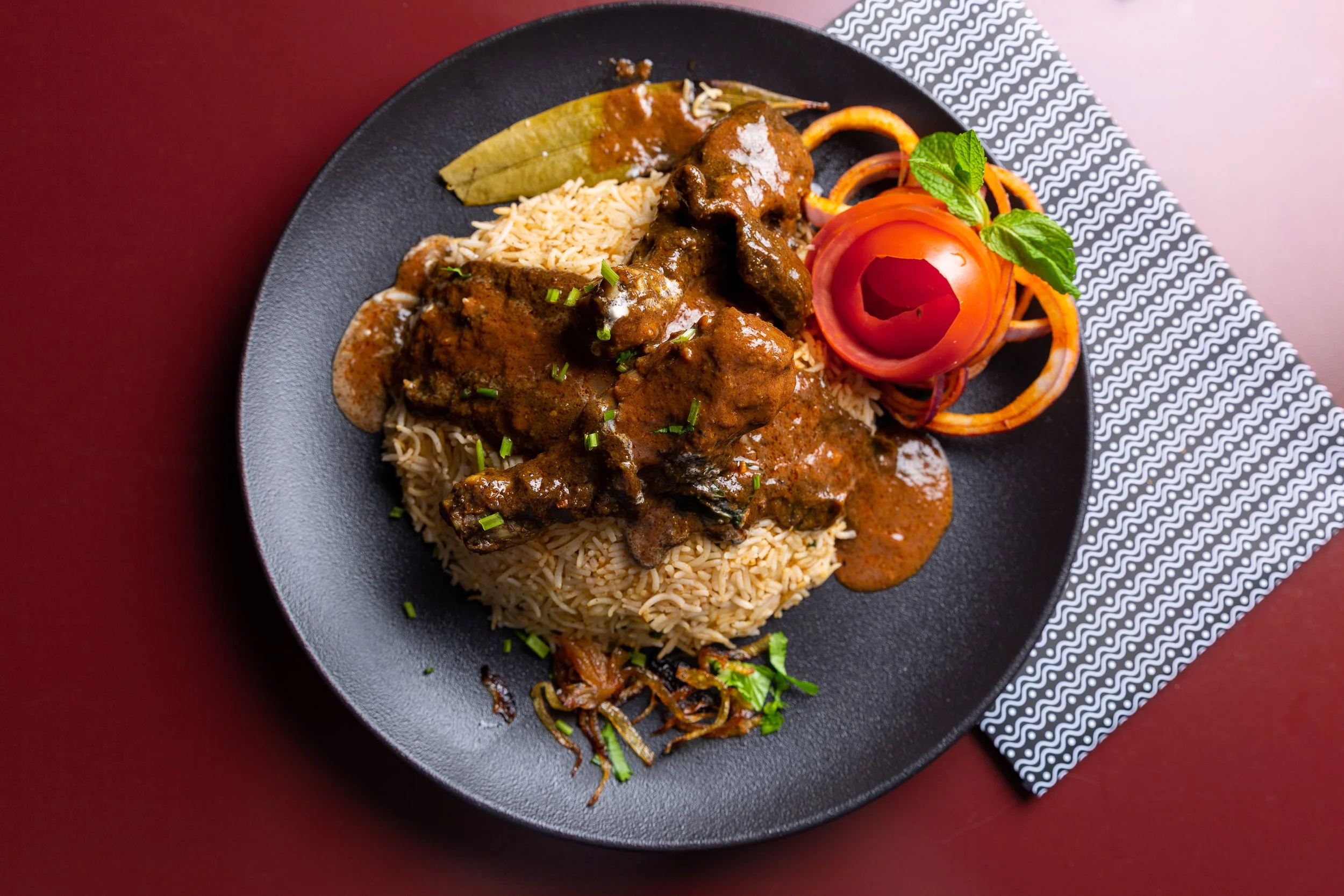 Plate of rice with curry, garnished with chopped green onions, and a side of vegetables including a tomato rose, lettuce, and sliced carrots, on black plate with patterned napkin.