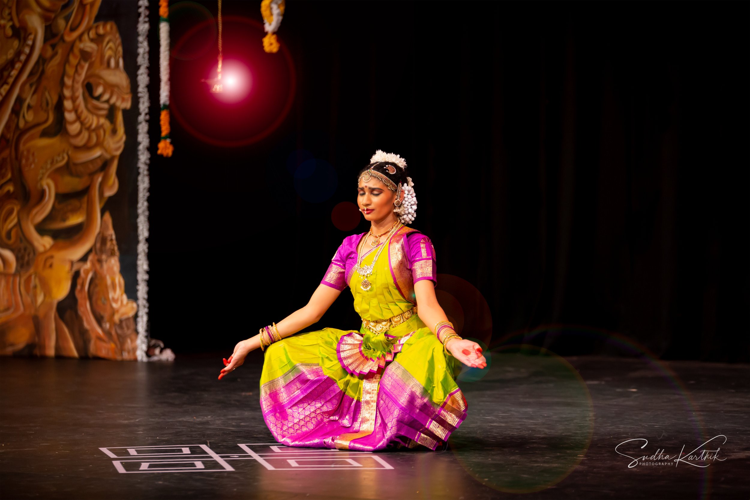 A woman dressed in traditional Indian attire performs a classical dance on stage. She is sitting on the floor with her legs crossed, wearing a bright green and pink sari, adorned with jewelry, flowers, and a headpiece. The stage background is dark wi