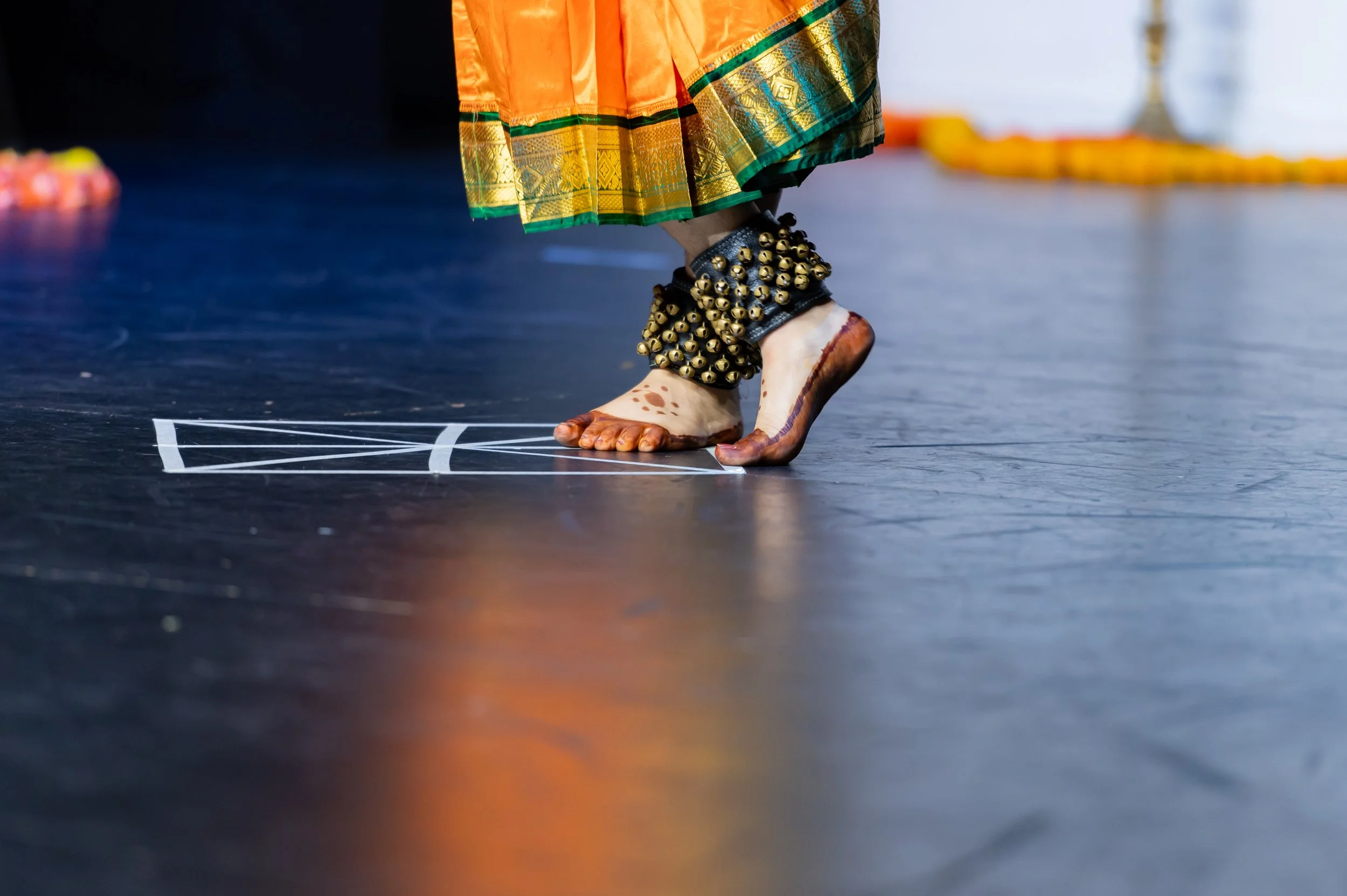 A person wearing traditional attire with ornate gold and green borders, anklets with bells, and henna designs on their feet, standing on a decorated floor during a festive occasion.