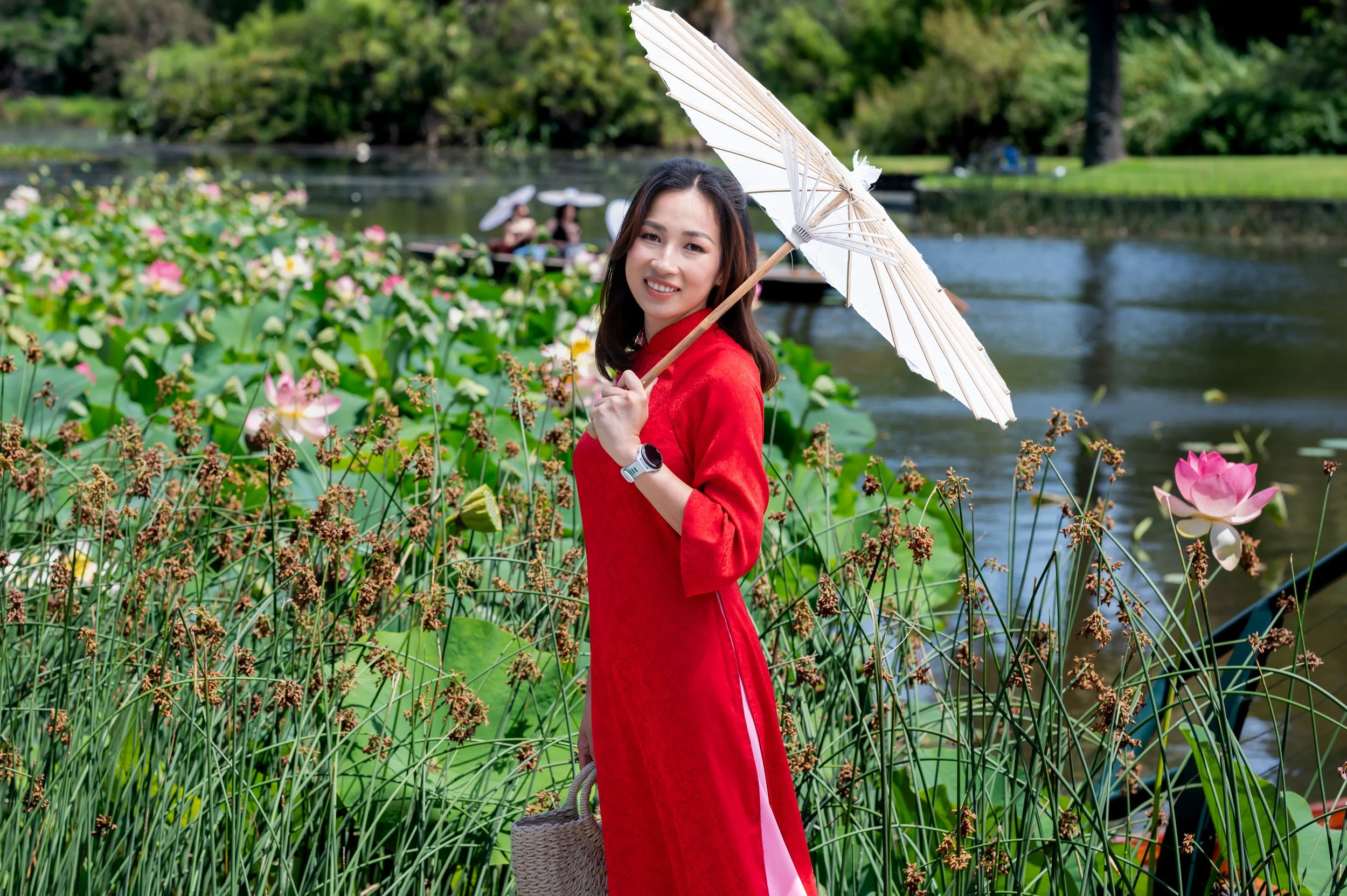A woman in a red traditional dress holding a white umbrella by a pond with lotus flowers and other people in the background.
