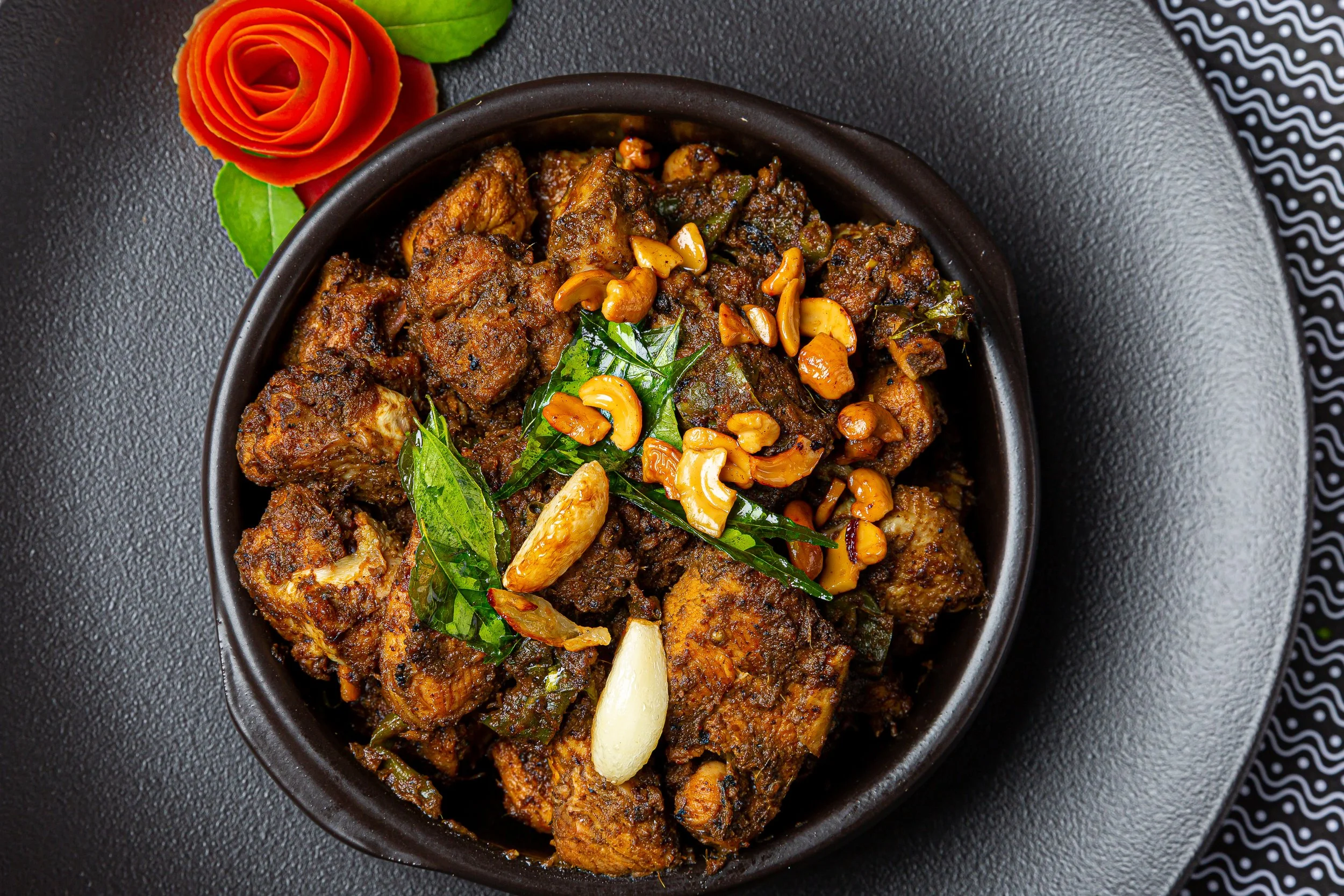 A black bowl filled with cooked Indian spiced meat, garnished with green herbs, cashews, and garlic, placed on a textured black surface with a decorative black and white patterned cloth nearby.