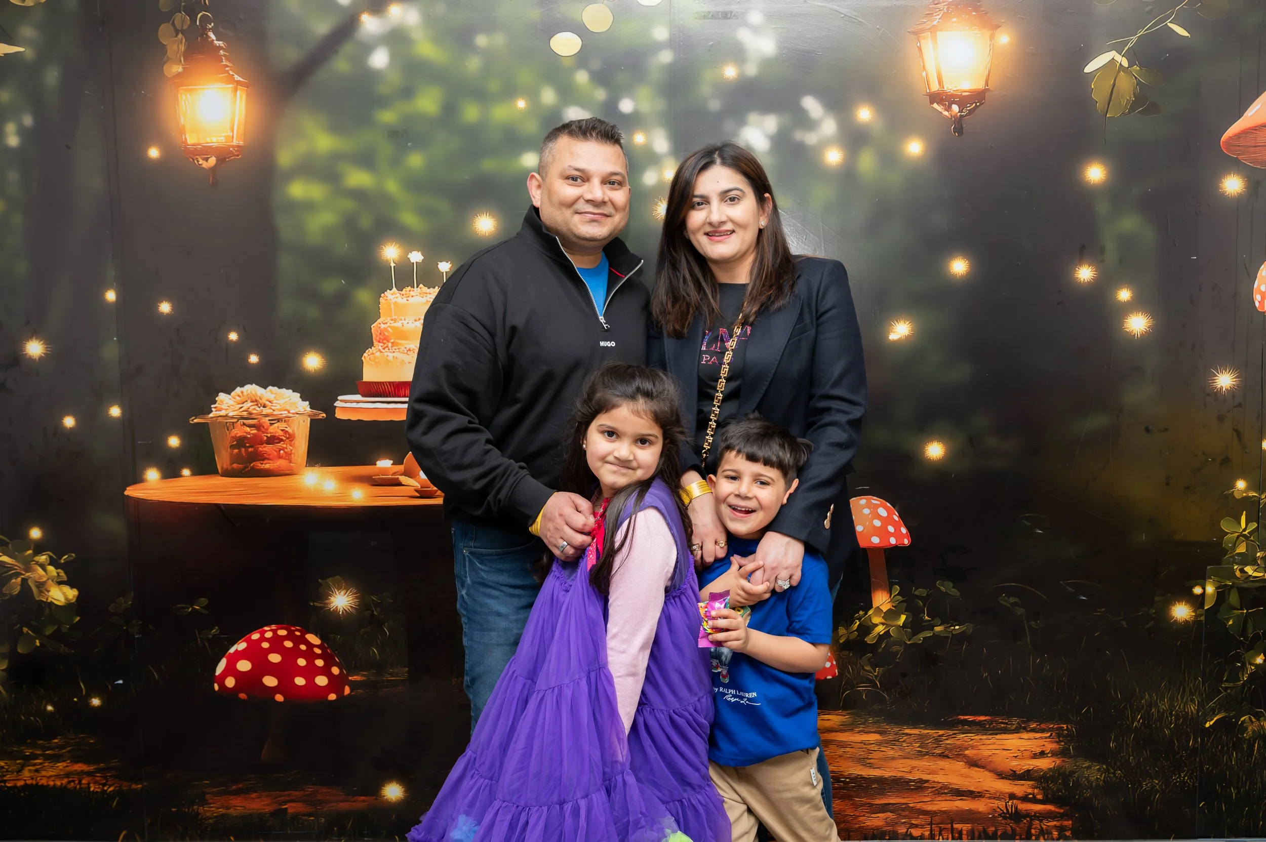 family of four, including a girl, a boy, a man and a woman, standing together at a birthday celebration with birthday cake, balloons, and glowing lights in the background.