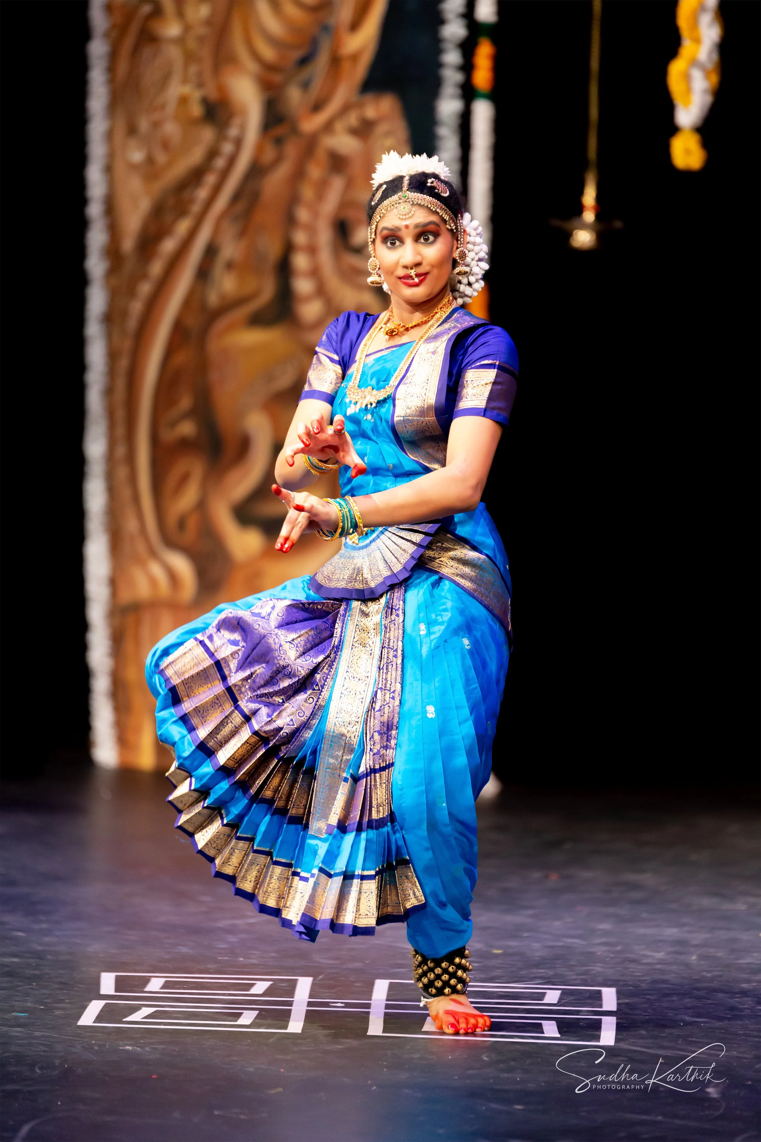 A woman dressed in traditional South Indian attire performing a classical dance on stage with a dark background and a wooden sculpture of a deity in the background.