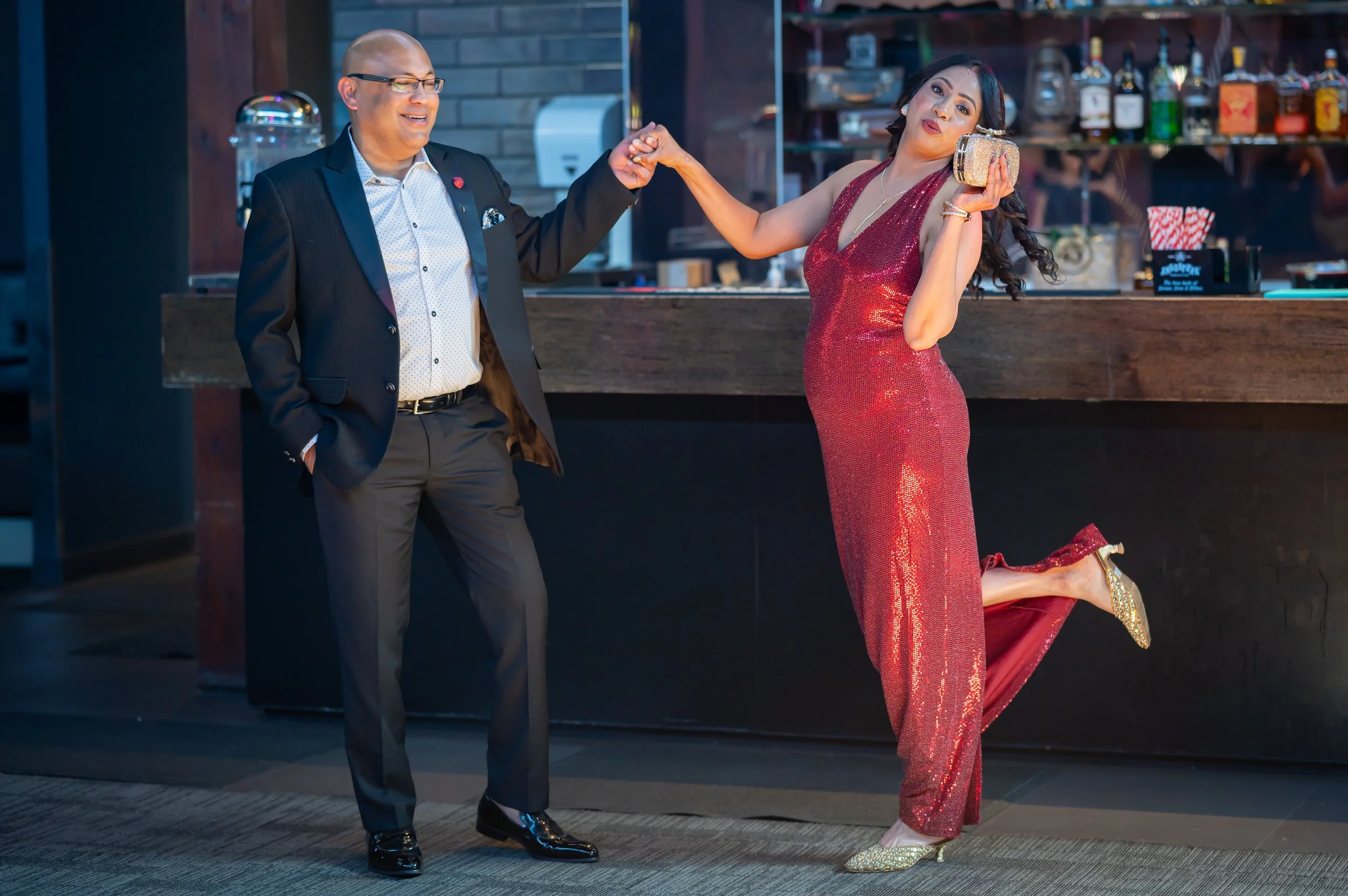 A man and a woman dancing together in a bar or club. The man is dressed in a dark suit with a white shirt, wearing glasses and smiling. The woman is wearing a sparkly red dress and holding a clutch purse, on her knees with one leg bent and the other 