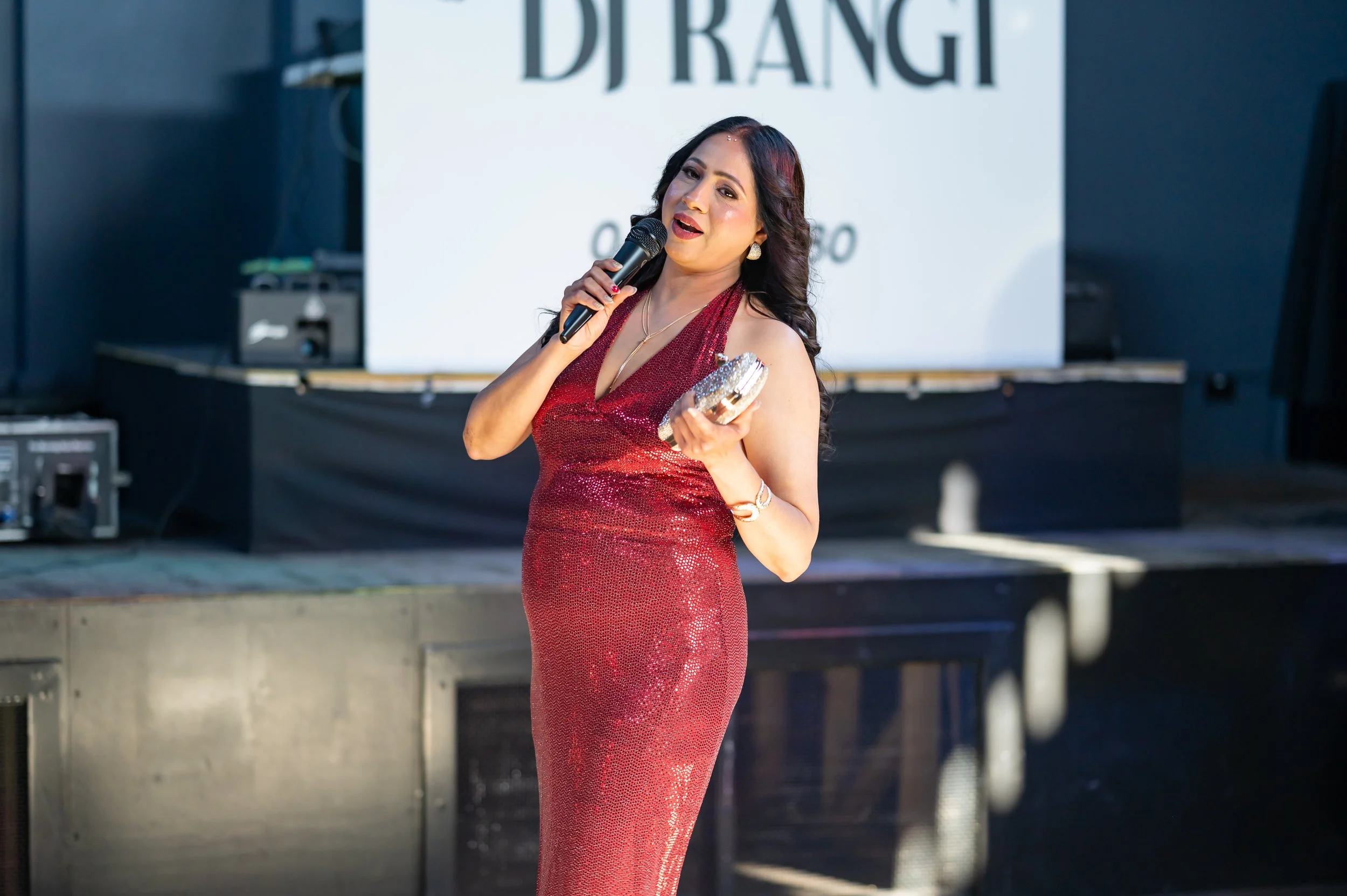 A woman in a red, sparkly evening gown speaking into a microphone on an outdoor stage during an event.