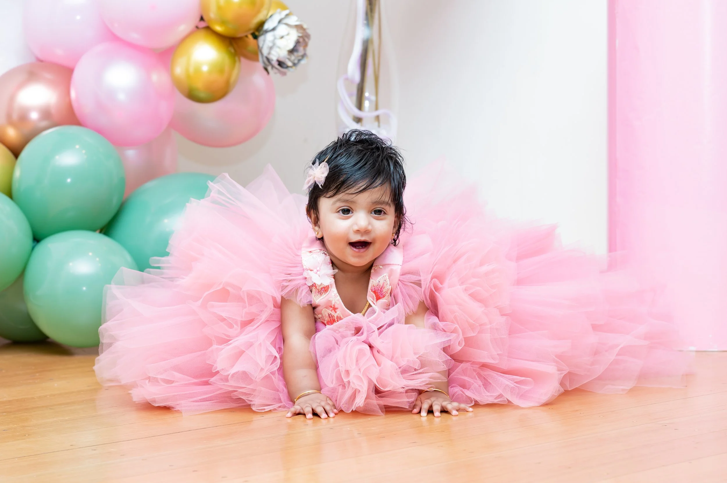 A young girl dressed in a pink tutu dress, smiling and crawling on a wooden floor, surrounded by pink and gold balloons for a celebration.