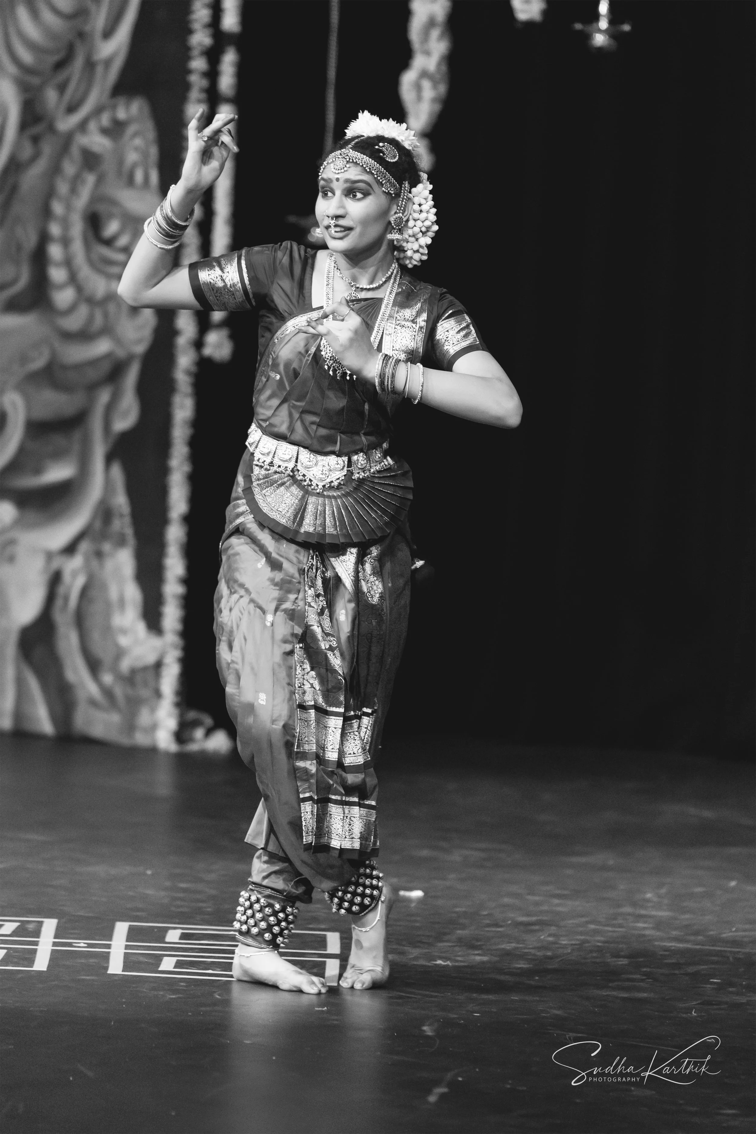 A woman in traditional Indian attire performing a dance on stage, wearing jewelry, a decorated blouse, and a sari, captured in black and white.