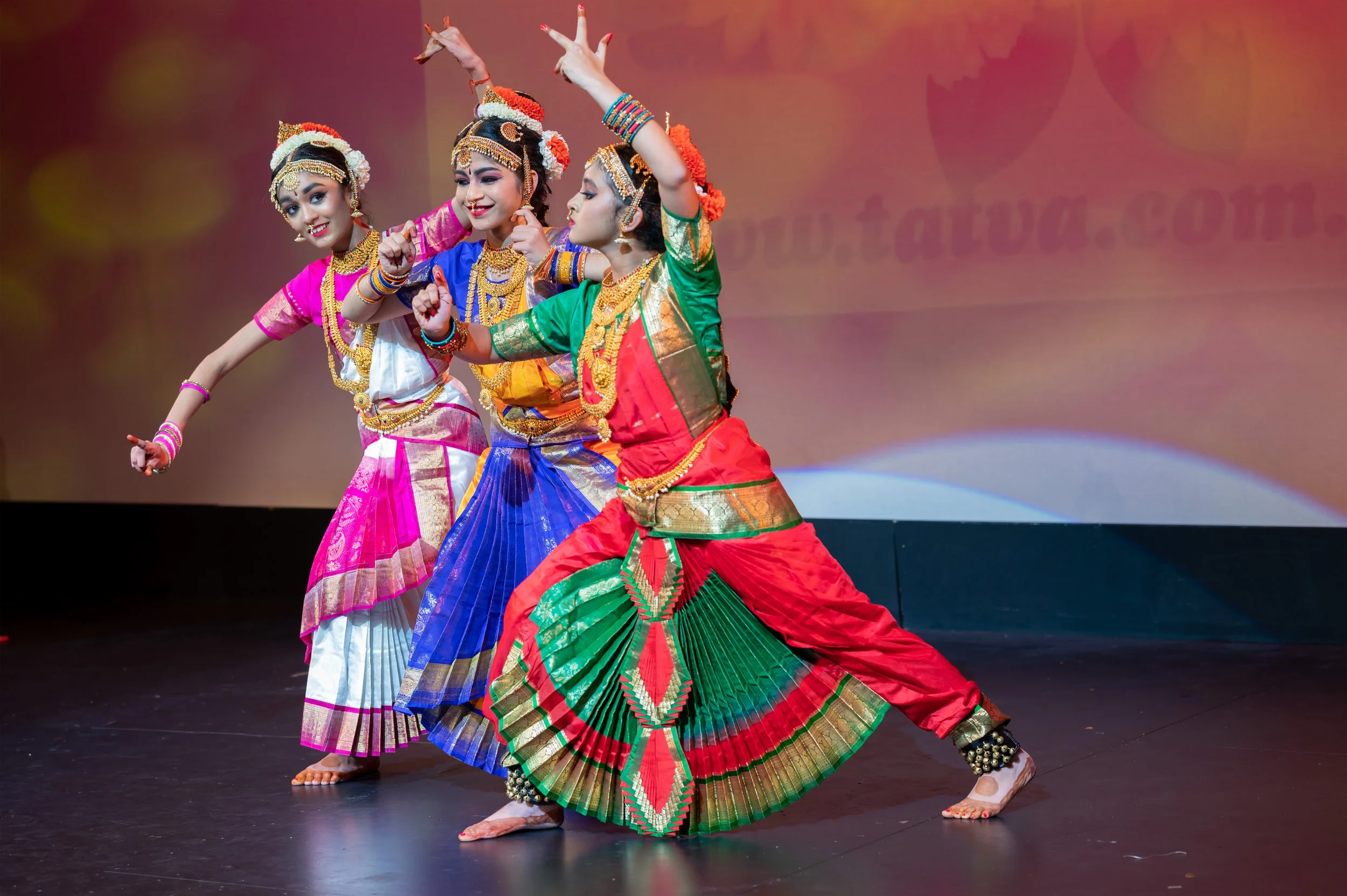 A group of women performing a traditional Indian dance on stage, dressed in colorful costumes with intricate jewelry and headpieces.
