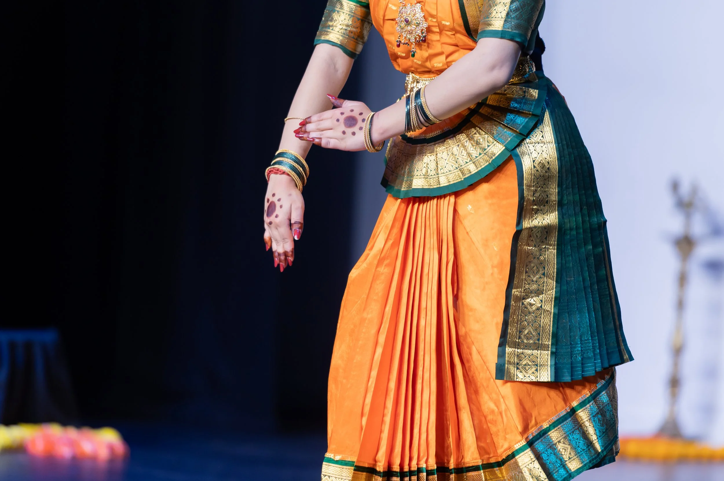 A person dressed in traditional Indian attire, wearing a colorful sari with gold accents, adorned with jewelry and bangles, holding their hands in a graceful pose