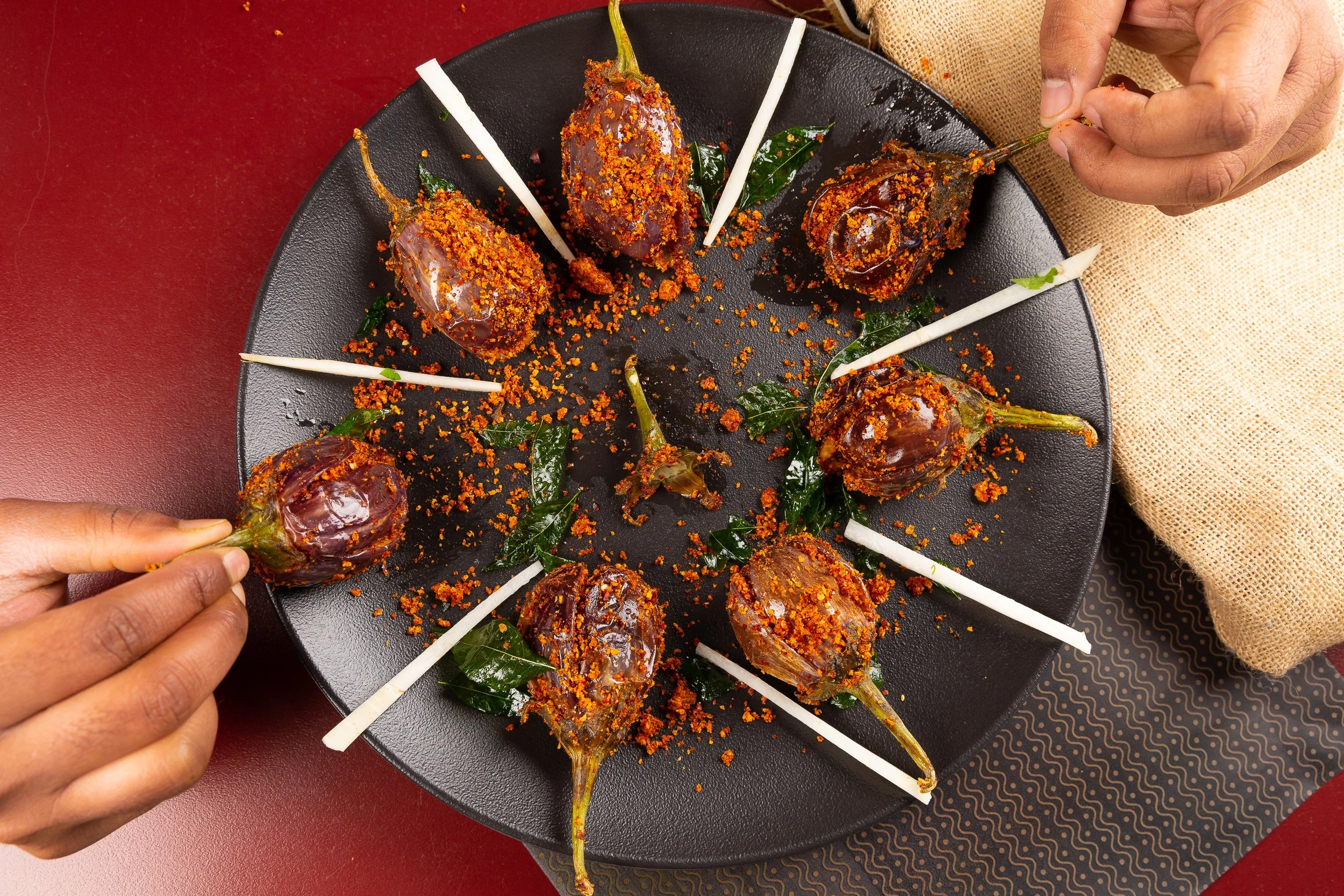 Person placing a roasted eggplant on a black plate, garnished with spices, green leaves, and white decorative sticks, on a table with a red surface and patterned placemat.