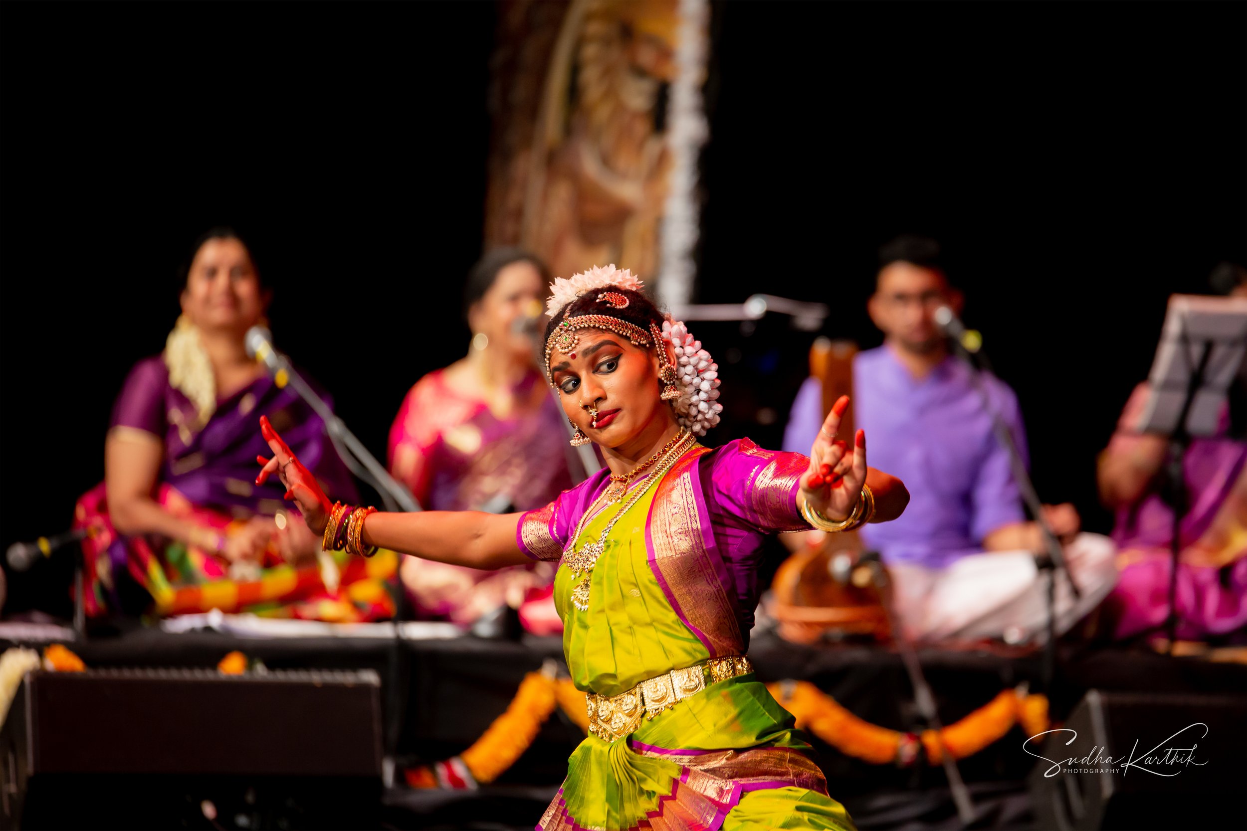 A woman dressed in traditional Indian attire performing classical dance on stage with a group of musicians in the background.