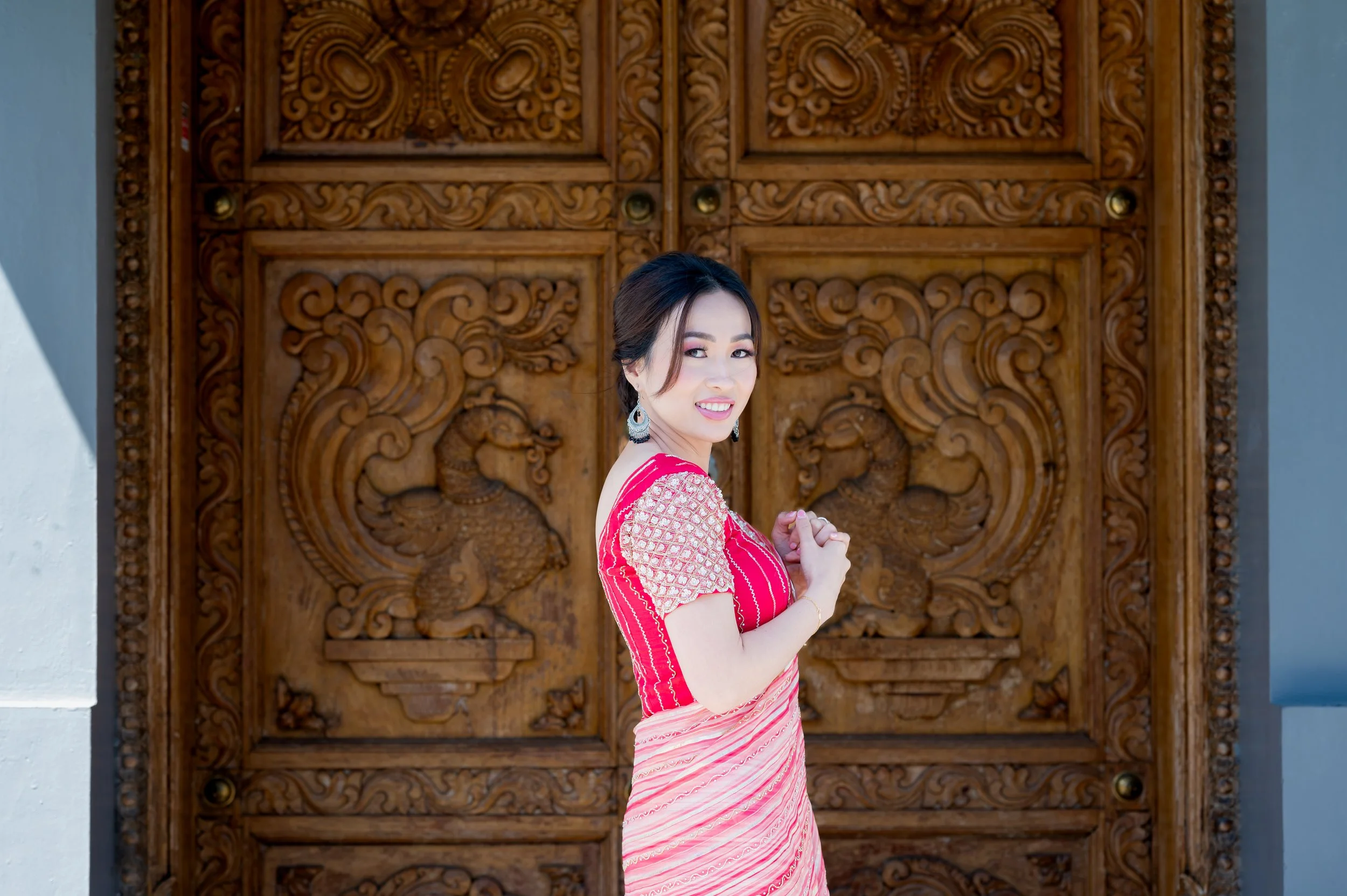 A woman dressed in a pink traditional outfit, smiling and standing in front of an ornately carved wooden door.
