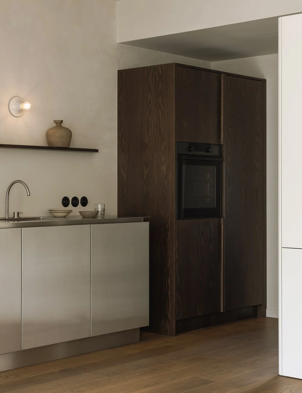Minimalistisk kjøkken i rustfritt stål med skap i mørk eik som tilfører varme og kontrast til det nedtonede interiøret.