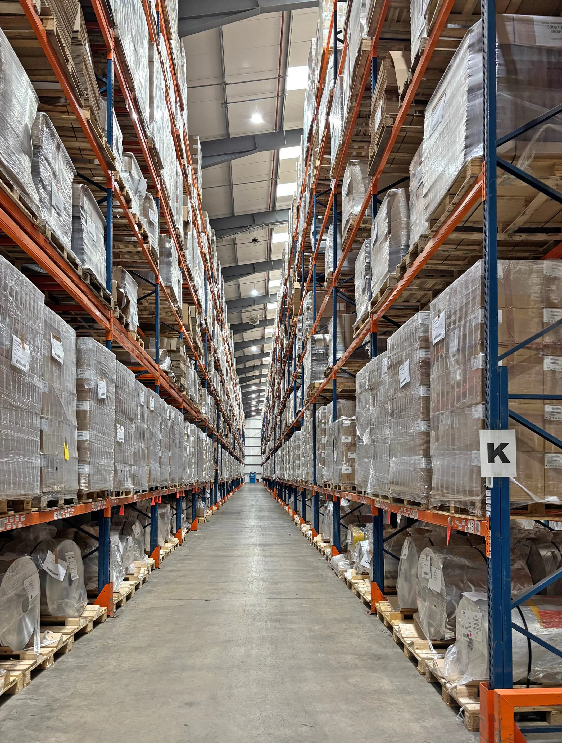 Long aisle in a warehouse with tall shelves filled with wrapped pallets of goods on each side, overhead lighting, concrete floor, and metal ceiling structure.