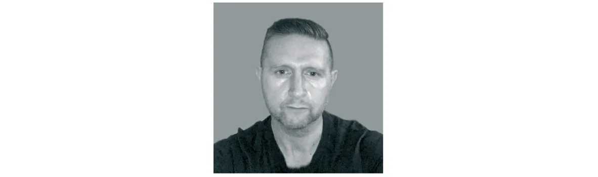 Black and white photo of a man with short hair, facial hair, wearing a dark shirt, against a plain background.