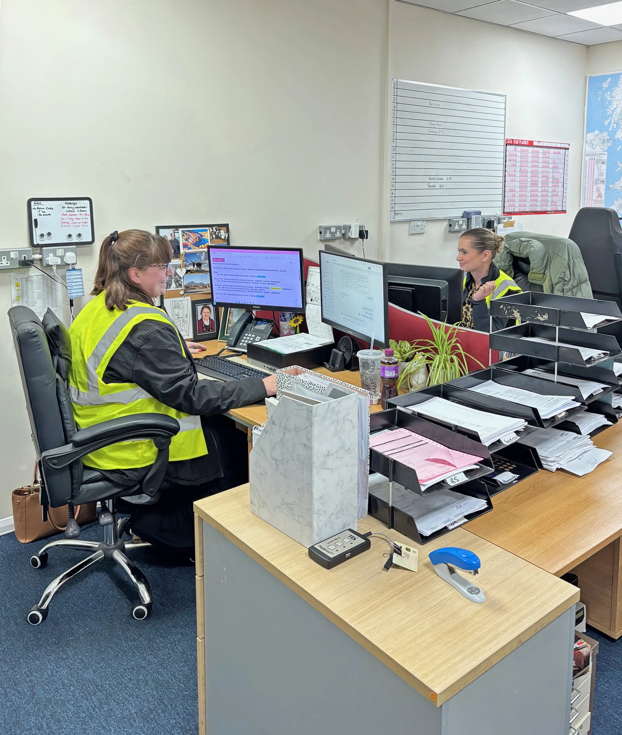 Office workspace with two women in bright yellow vests working at desks with computer monitors, surrounded by office supplies, plants, and paperwork, in a professional environment.