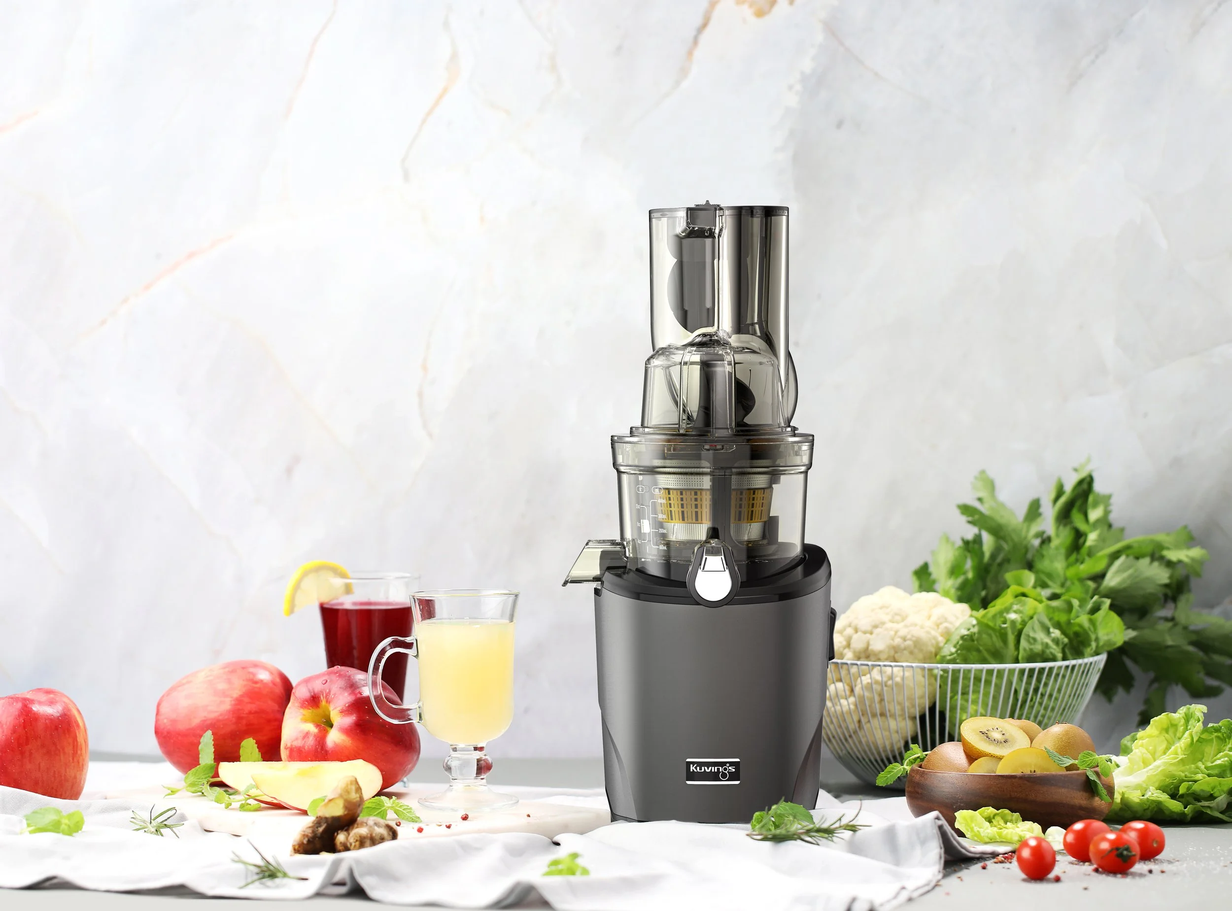 A Kuvings juicer on a table with apples, tomatoes, broccoli, cauliflower, kiwi, and ingredients for juice and vegetables in bowls and on the table.