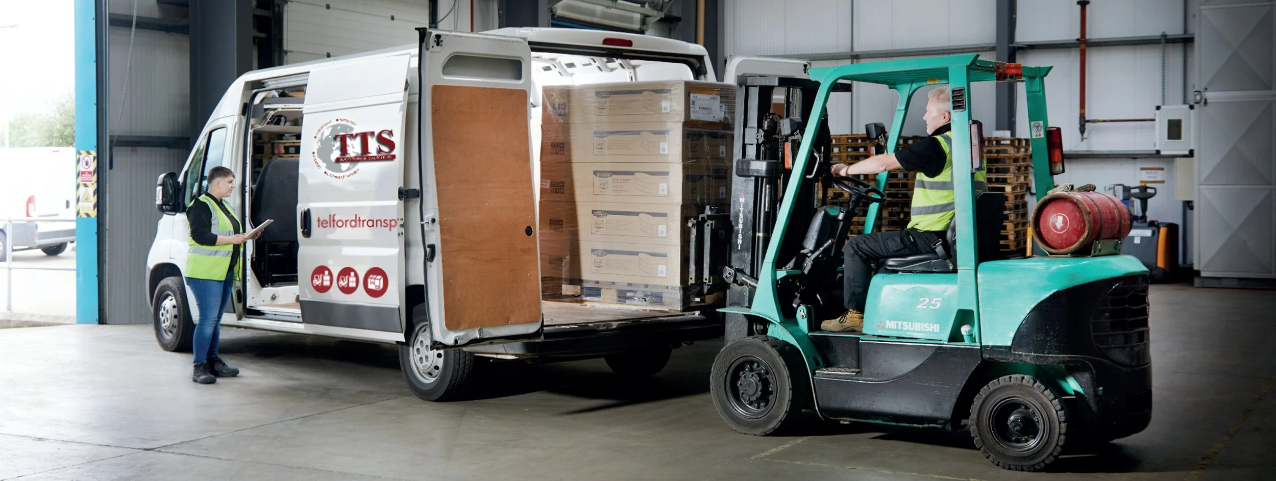 A worker operating a green forklift to load boxes into a white delivery van inside a warehouse. A woman wearing a yellow safety vest and holding a clipboard is standing nearby."
