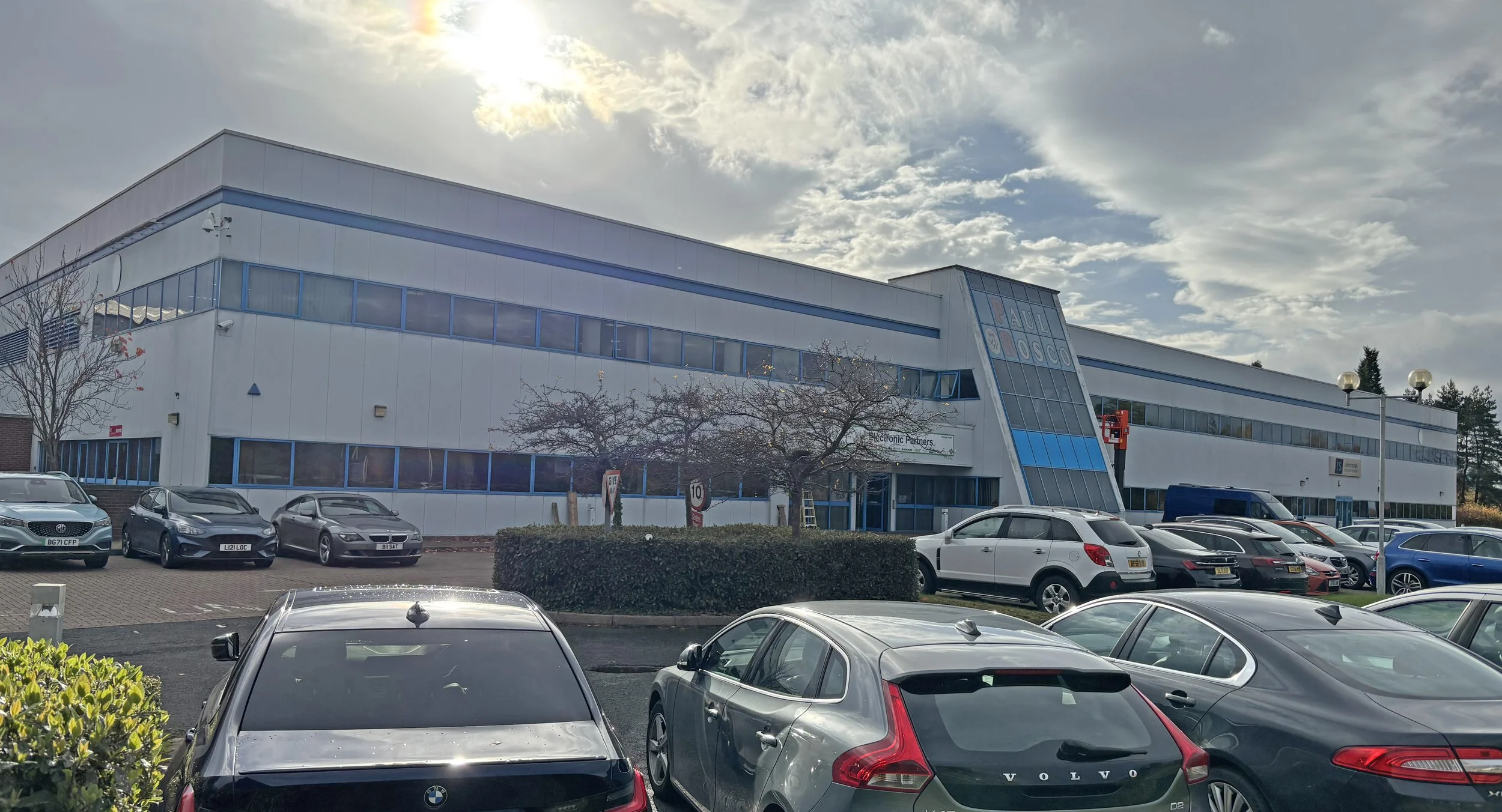 Large commercial building with multiple windows, blue accents, and a sign that reads 'PAUL OSCO'. The parking lot in front has various cars, and a clear sky with partly cloudy weather is visible.
