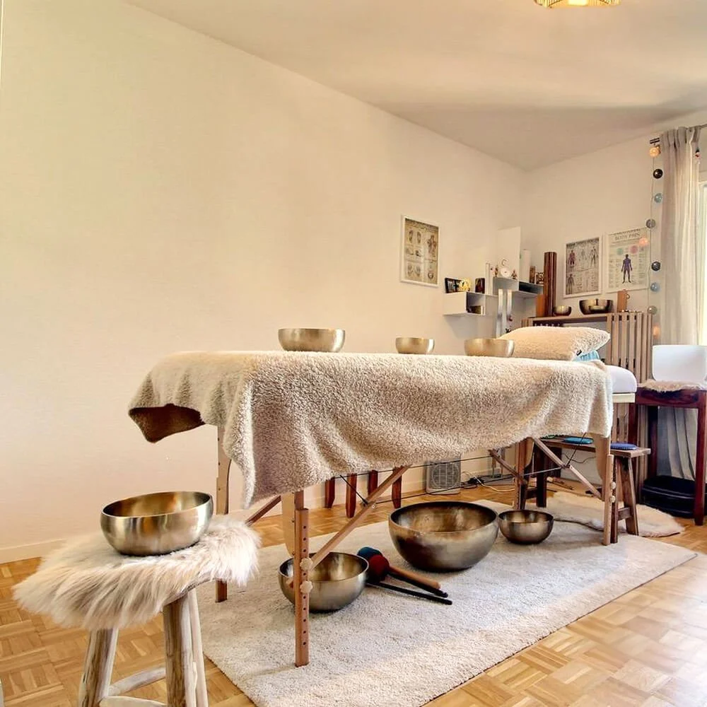 Une table de massage recouverte d'une serviette beige, avec plusieurs bols en métal posés dessus, dans une pièce bien éclairée avec un tapis et des éléments de décoration sur un mur.