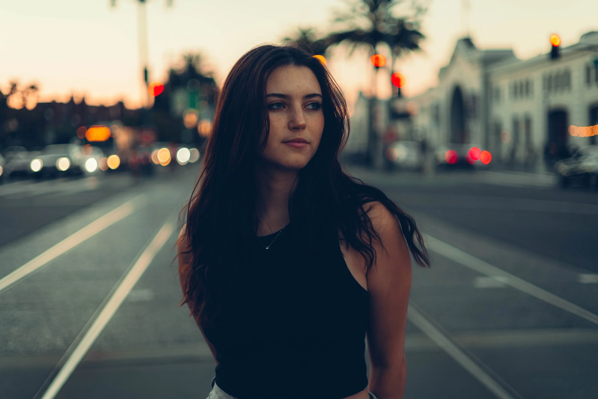 A young woman with long dark hair, wearing a black sleeveless top, stands on city tram tracks during sunset or dusk with blurred city buildings and palm trees in the background.