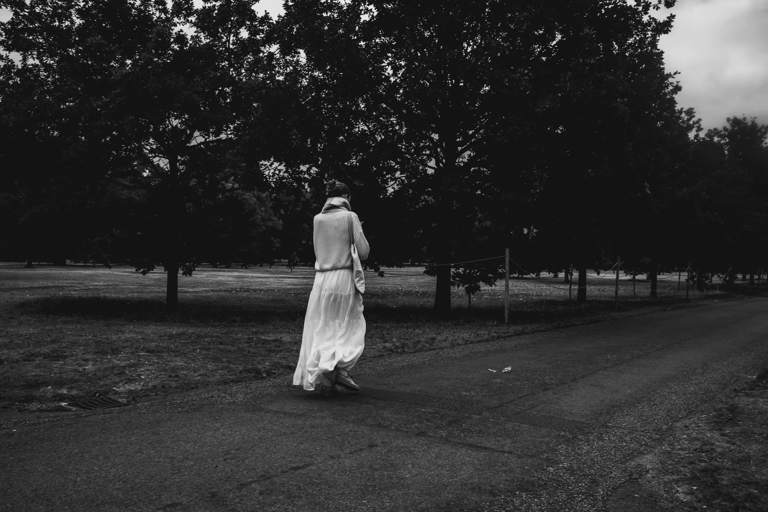 A woman in a long white skirt and sweater walking alone on a path near trees in a park, captured in black and white.