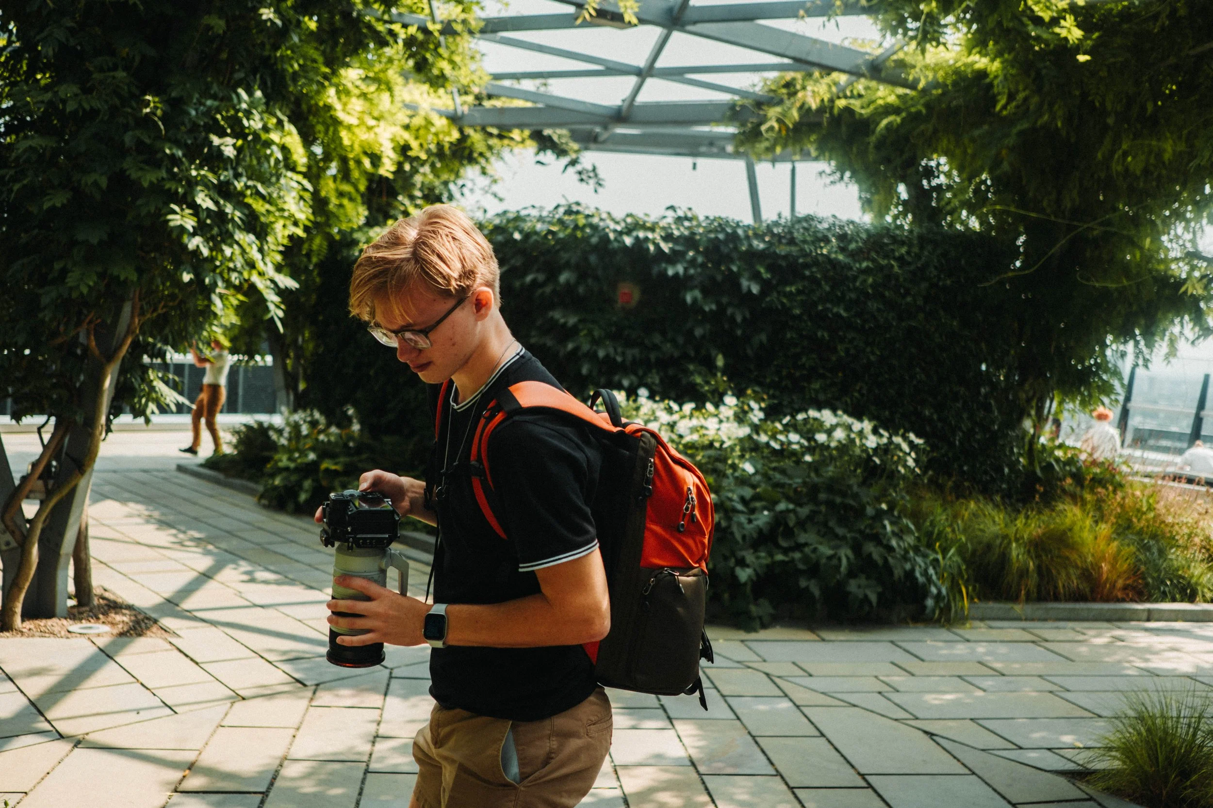 Young man in black T-shirt and khaki shorts holding a camera with a lens, wearing glasses and a smartwatch, carrying a red and black backpack, walking outdoors on a sunny day in a green, shaded garden area.