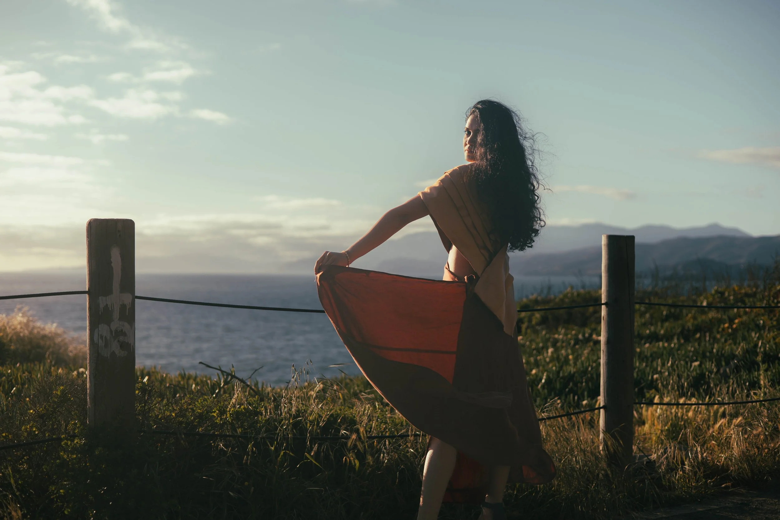 A woman standing outdoors near the ocean during sunset, holding her dress, with mountains in the background.