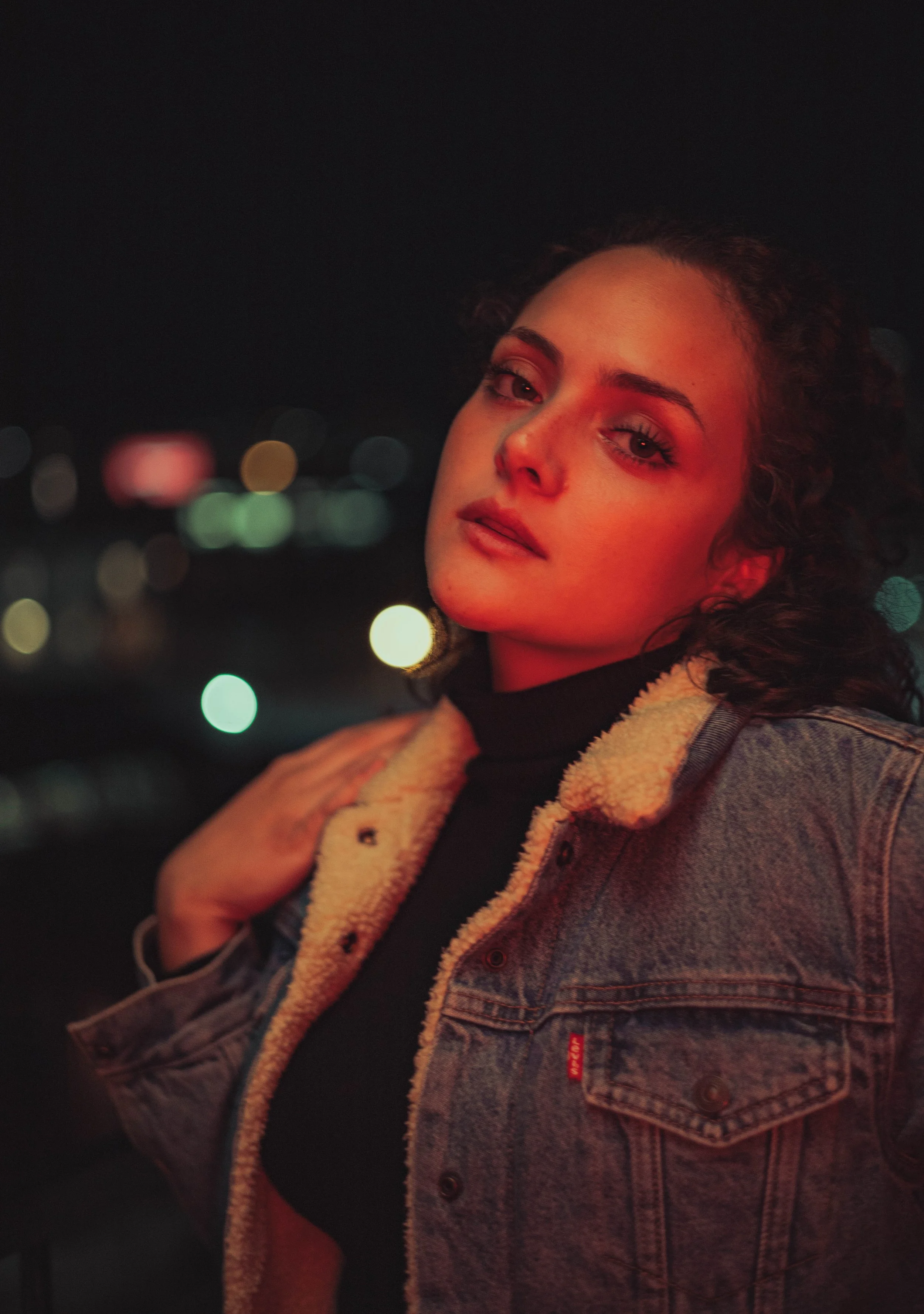 A young woman with curly hair and makeup posing at night with city lights in the background, wearing a denim jacket with a shearling collar.