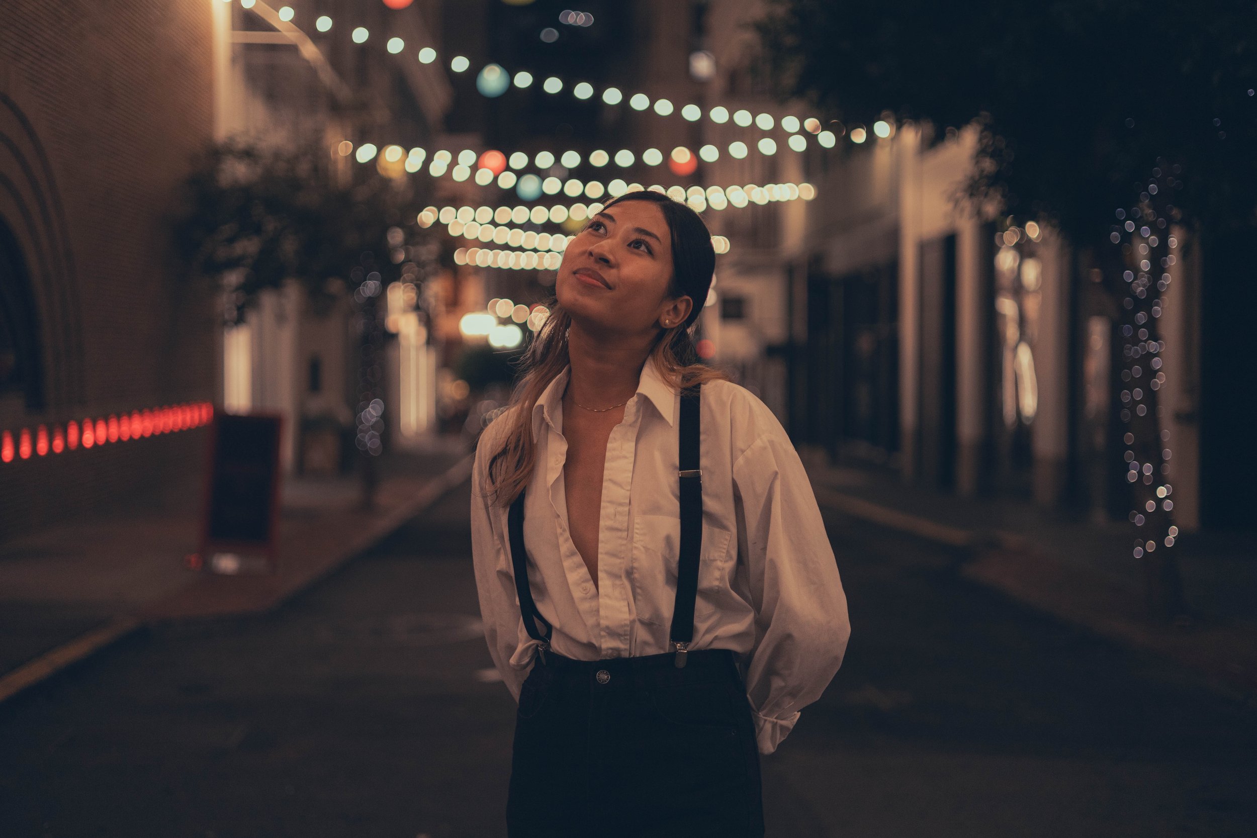A woman standing on a city street at night, looking up with a thoughtful expression. The street is decorated with string lights, creating a warm and festive atmosphere.