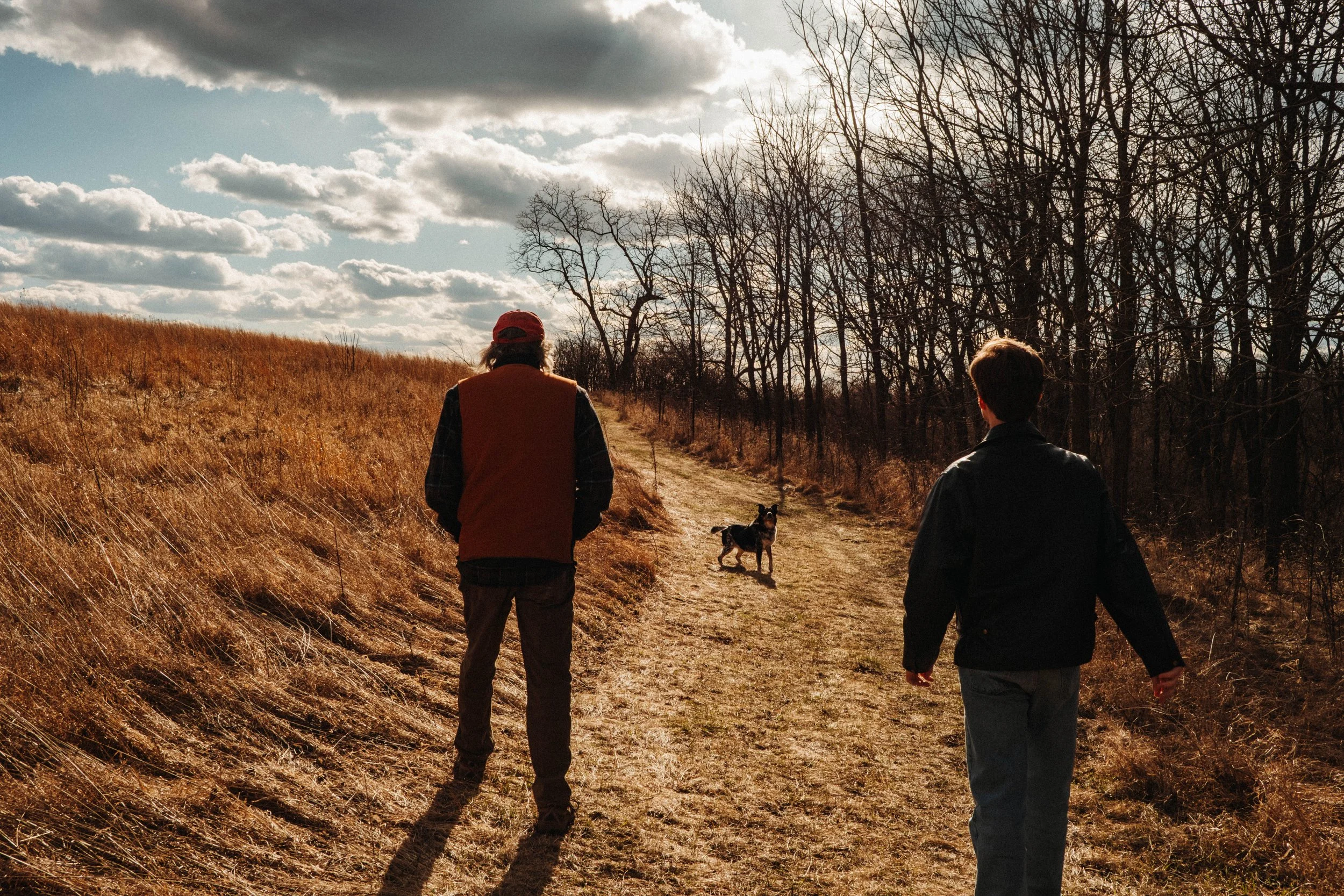 Two men, one with a red vest and a cap, and the other with a black jacket, walk along a dirt trail in a rural landscape during late fall, with a dog running ahead. The scene features tall, dry grass on one side and leafless trees on the other, under 