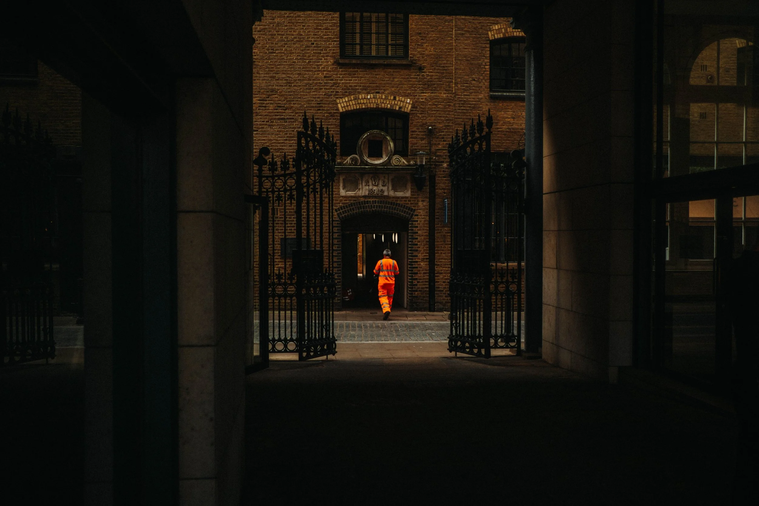 Person in orange reflective safety vest and pants walking through a gated alley towards a brick building.