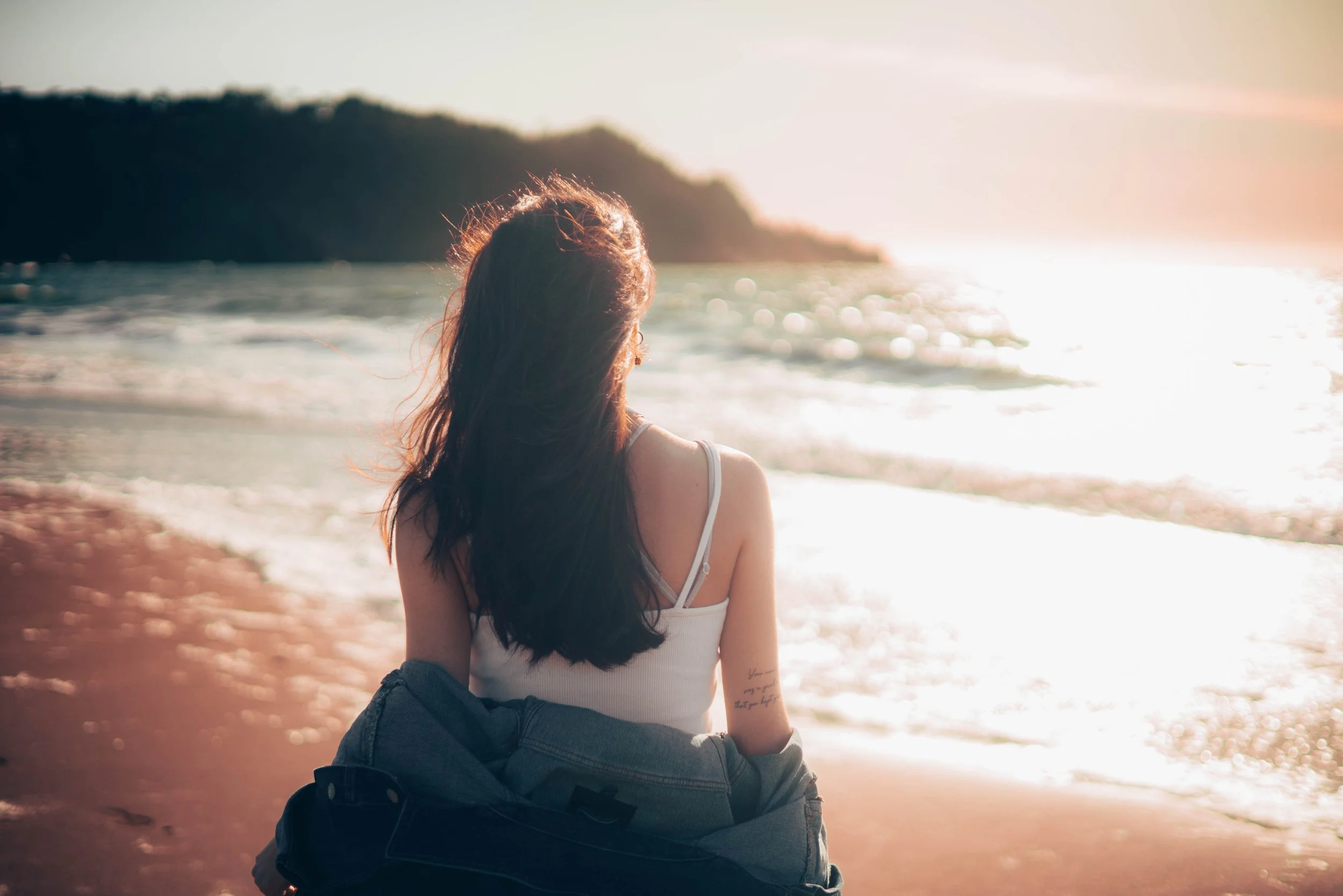 A woman with dark hair sitting on the beach at sunset, facing the ocean, wearing a white tank top and a denim jacket.