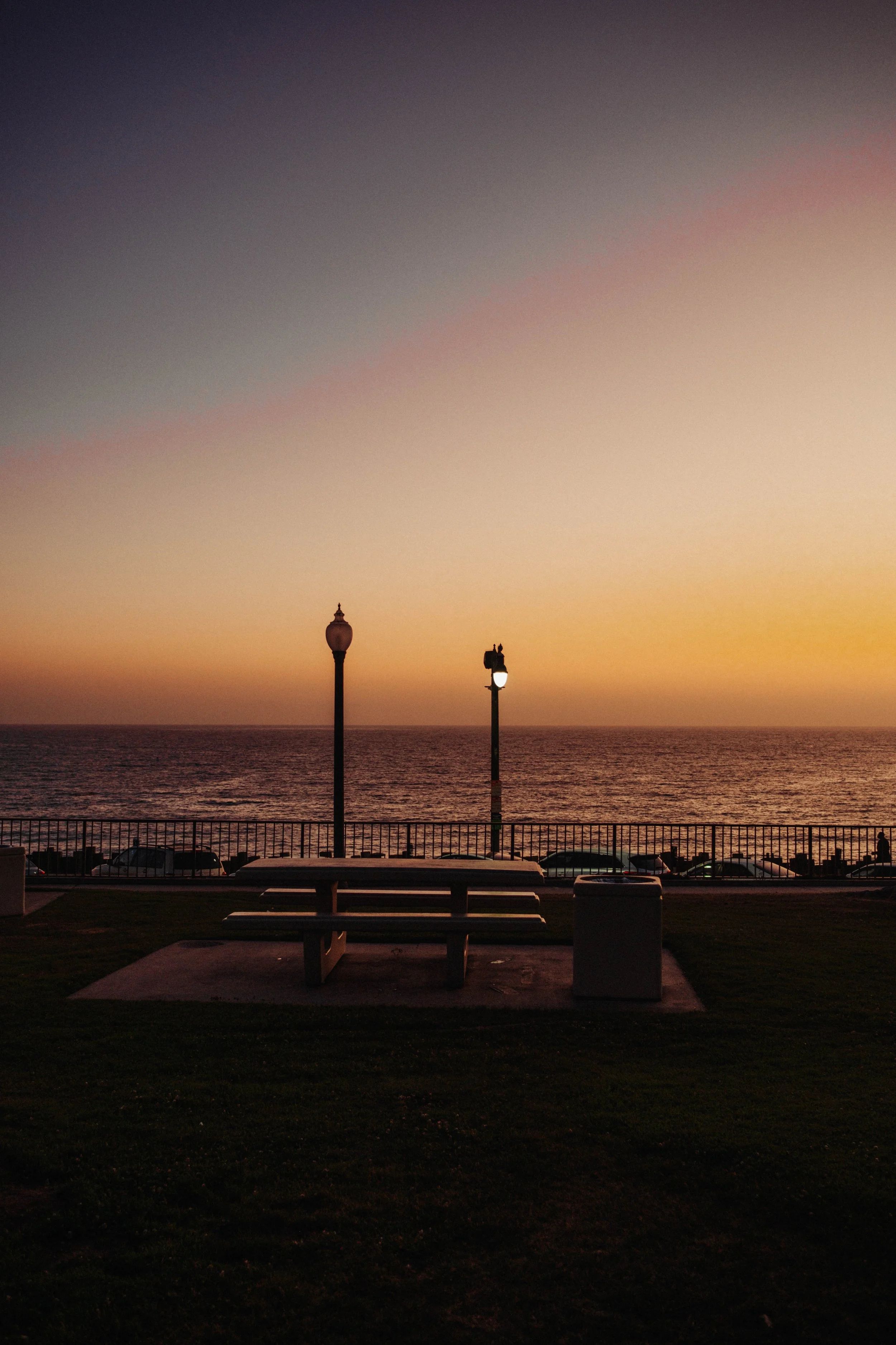 A park bench with a trash can in front of a fence overlooking the ocean at sunset. Two street lamps are visible, one with a decorative top and the other with a simple bulb. The sky displays a gradient of colors from orange near the horizon to dark bl