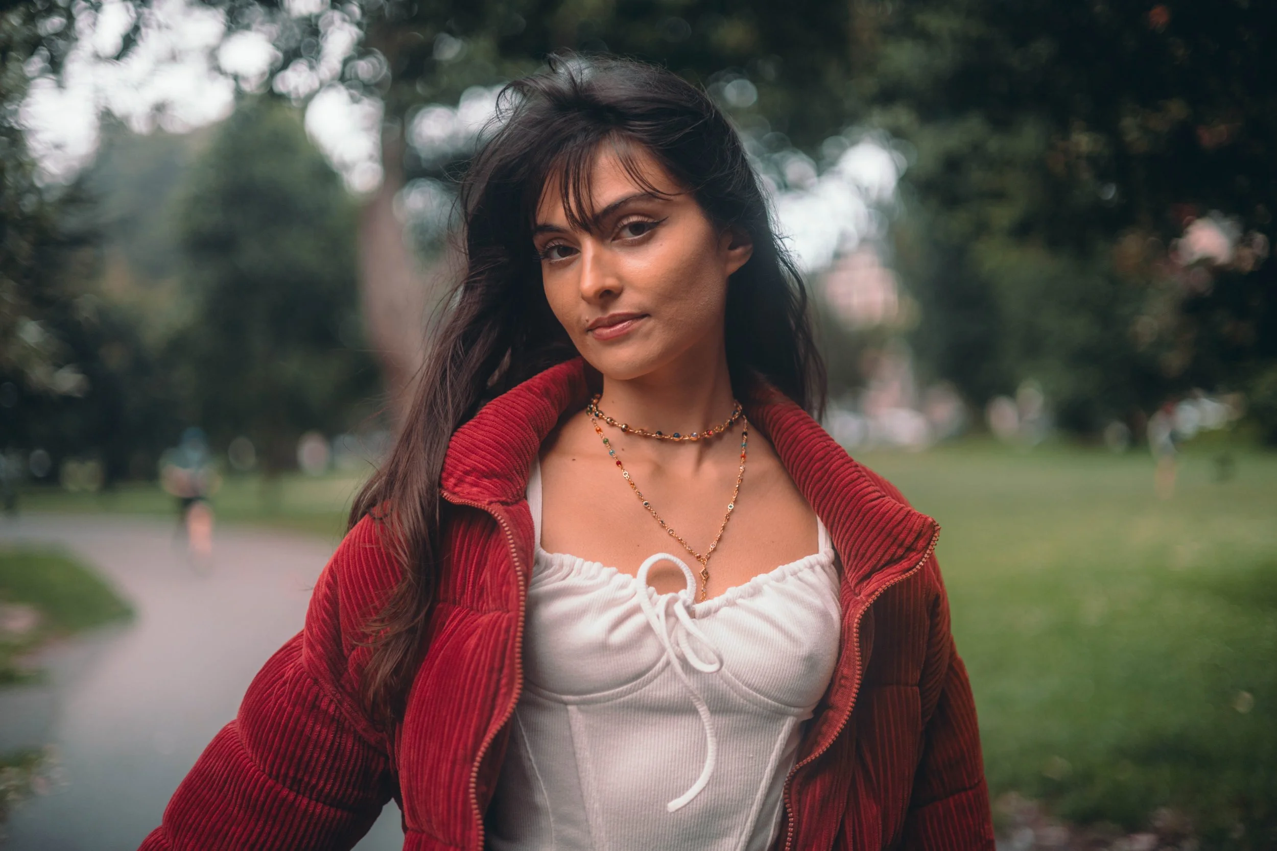A woman with long dark hair standing outdoors in a park-like setting, wearing a red jacket over a white top and layered necklaces. There are trees and a grassy area in the background.