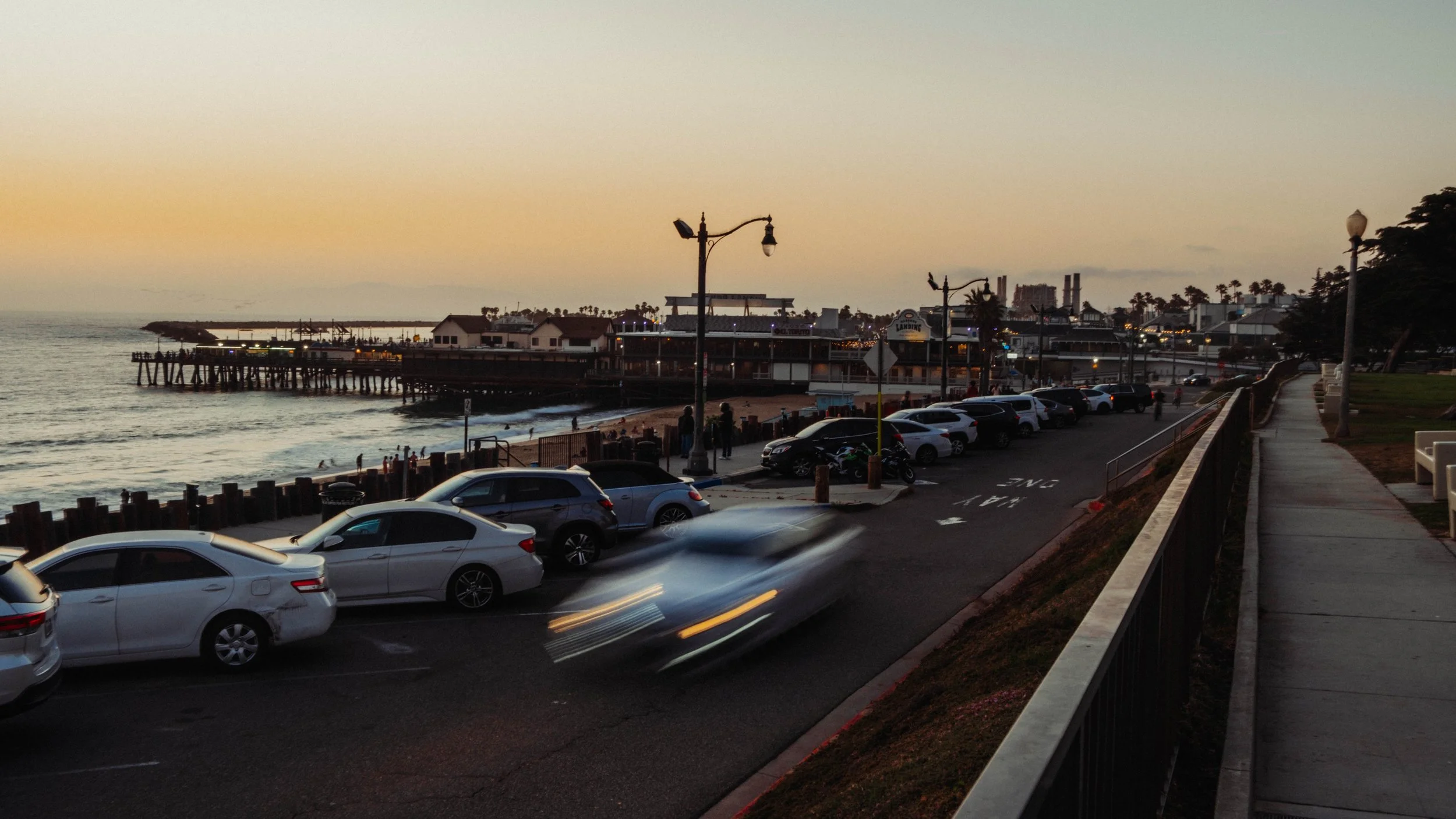 Beachside parking lot at sunset with cars, a pier extending into the ocean, and a boardwalk with street lamps.